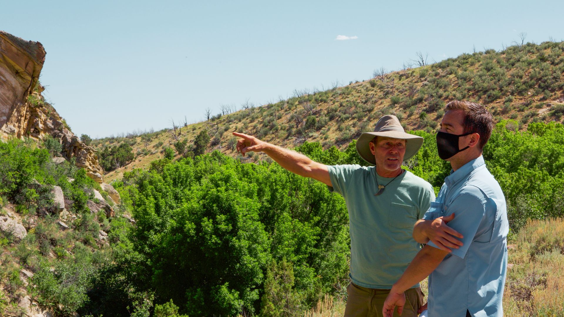 A man in a wide-brim hat points toward a cliffside while speaking to another man wearing a face mask at Blind Frog Ranch.