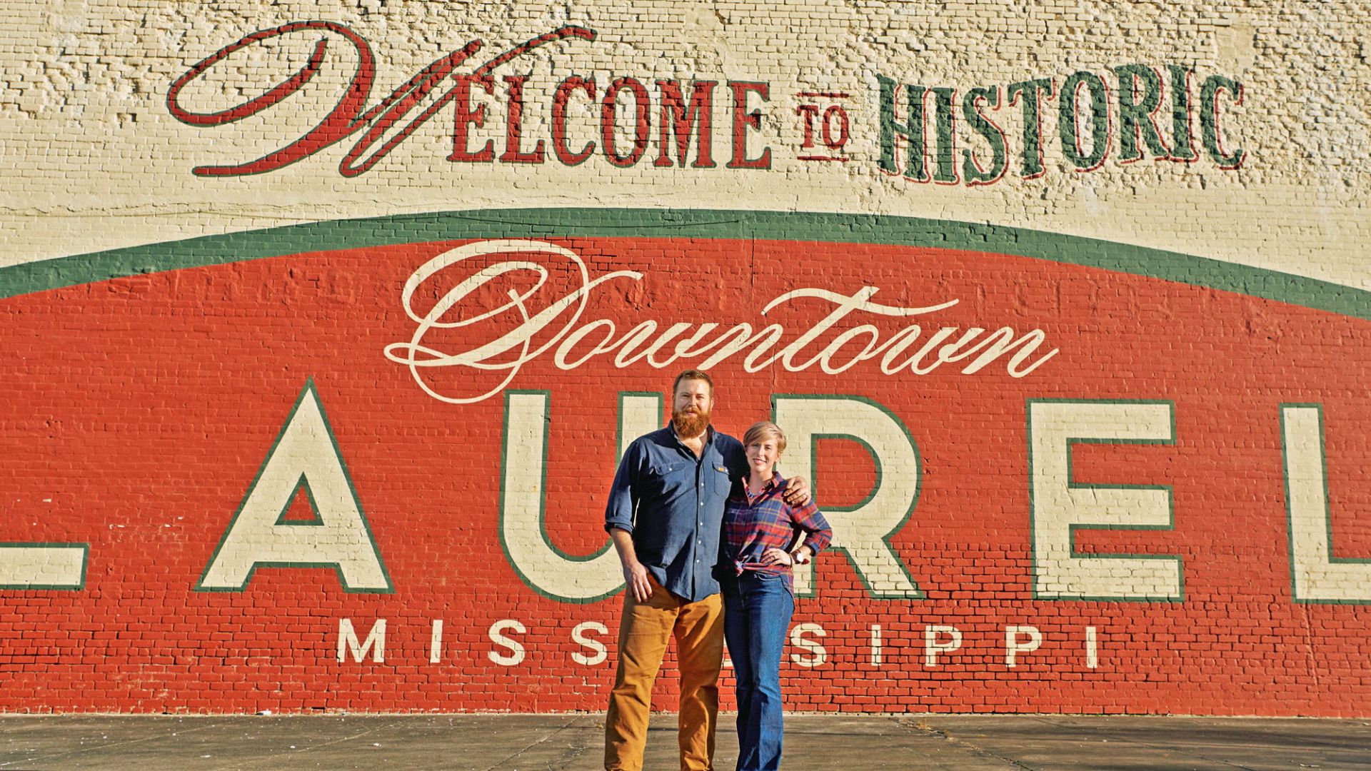 Ben and Erin Napier of HGTV’s Home Town posing in front of the red "Welcome to Historic Downtown LAUREL" mural.