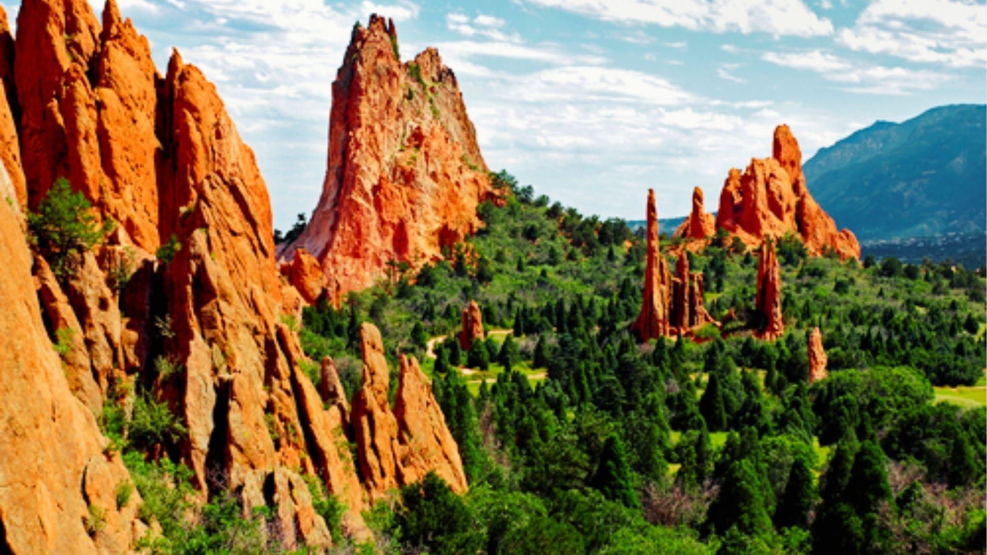 Towering red sandstone rock formations rise above green pine forest under partly cloudy blue sky