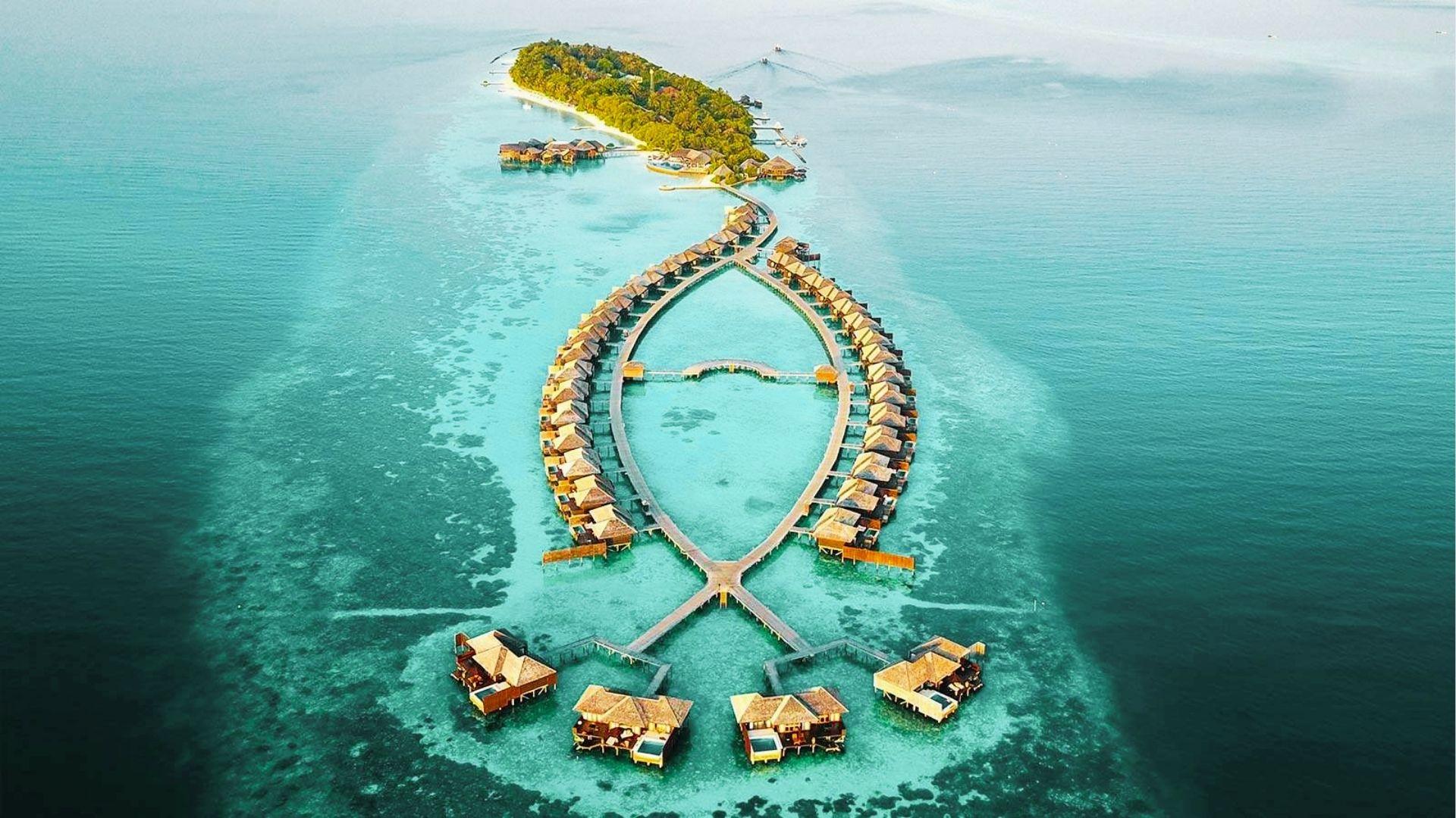 Aerial of overwater villas arranged in looped pattern around turquoise lagoon by lush island resort