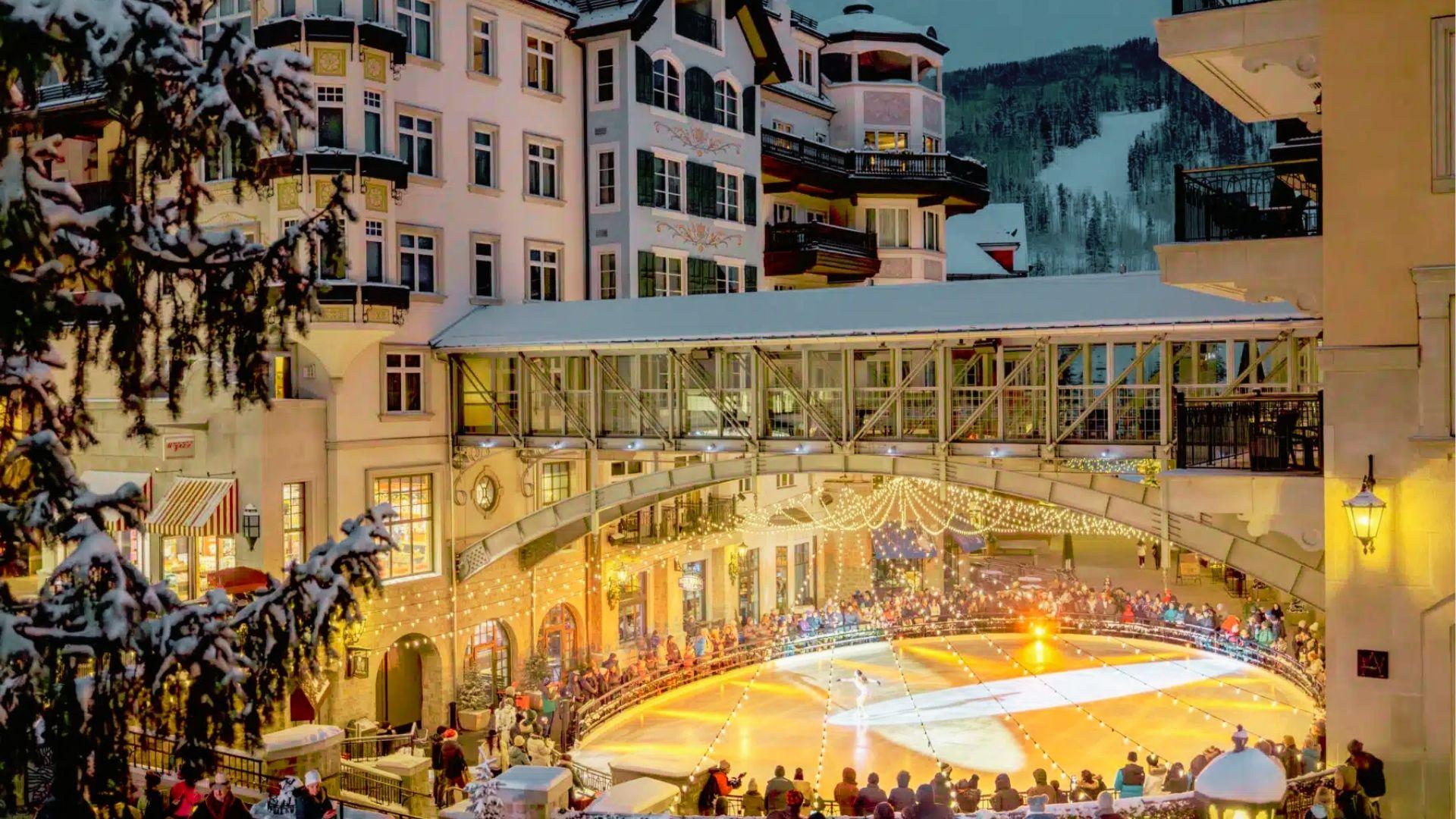 Evening alpine village with lit ice rink, skaters, snow covered buildings, and ski slope behind