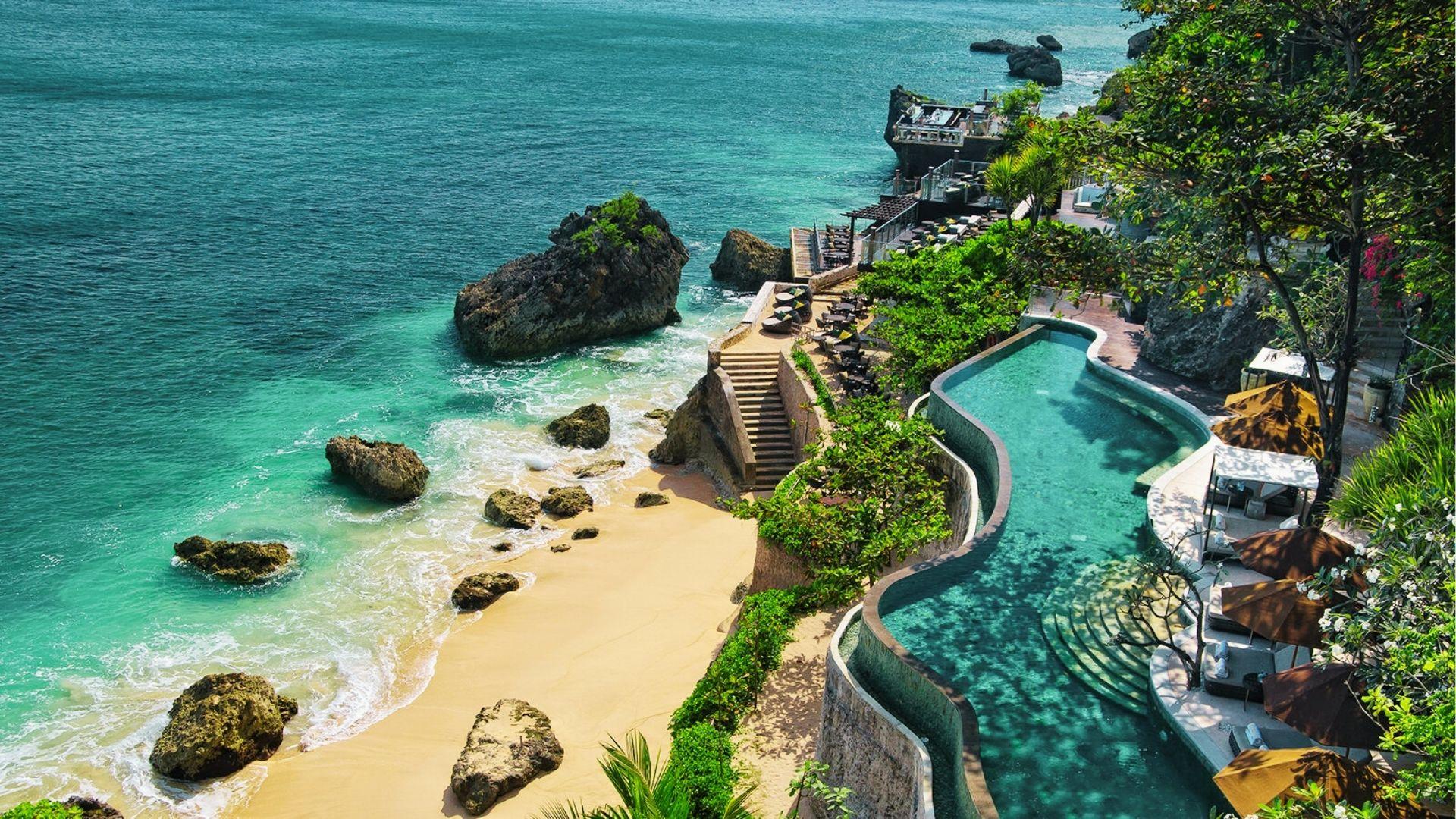 Cliffside infinity pool overlooks sandy beach, ocean rocks, turquoise water with lounge umbrellas below