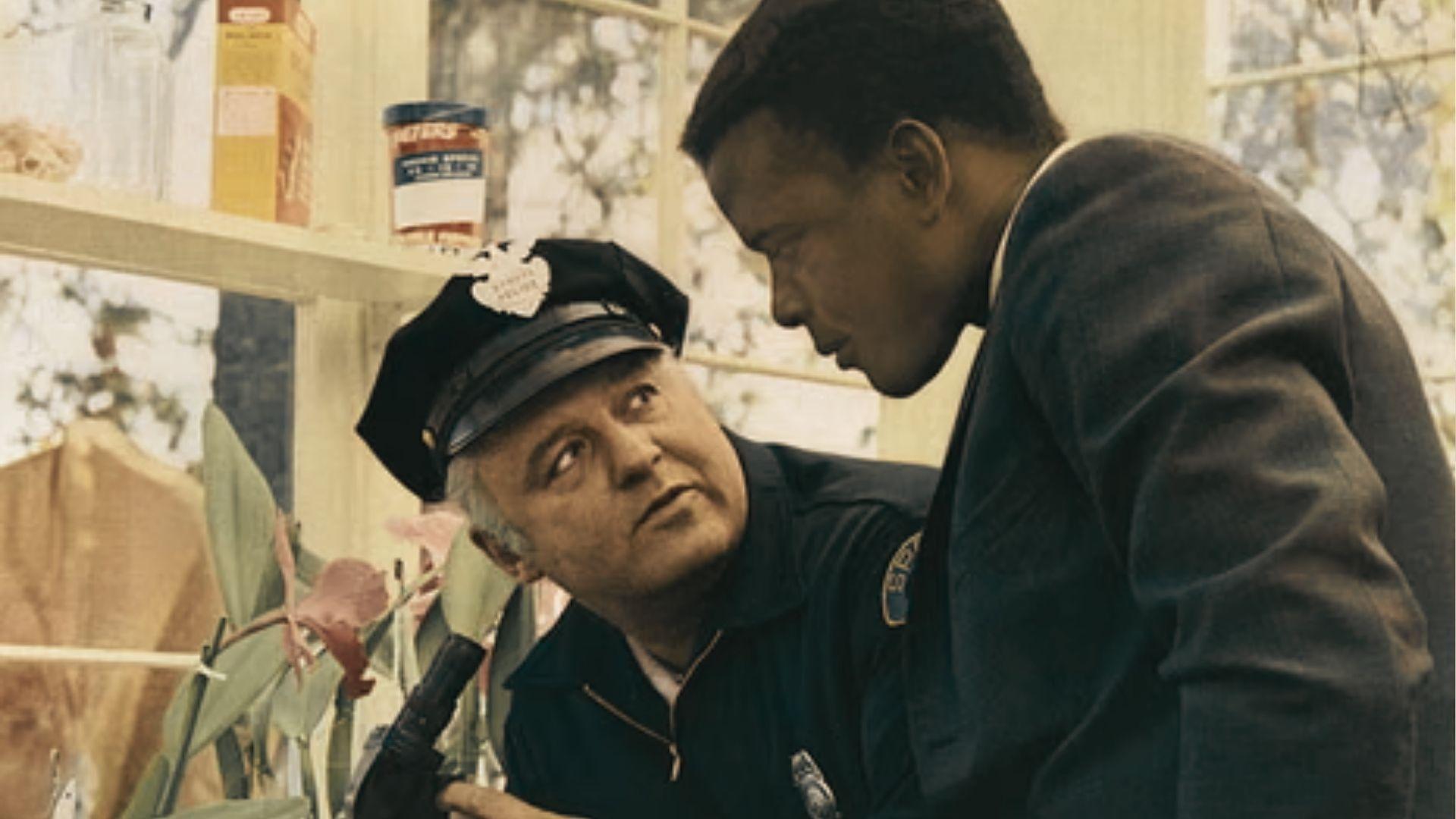 Sidney Poitier as Virgil Tibbs leans over to speak with a uniformed police officer in a greenhouse.