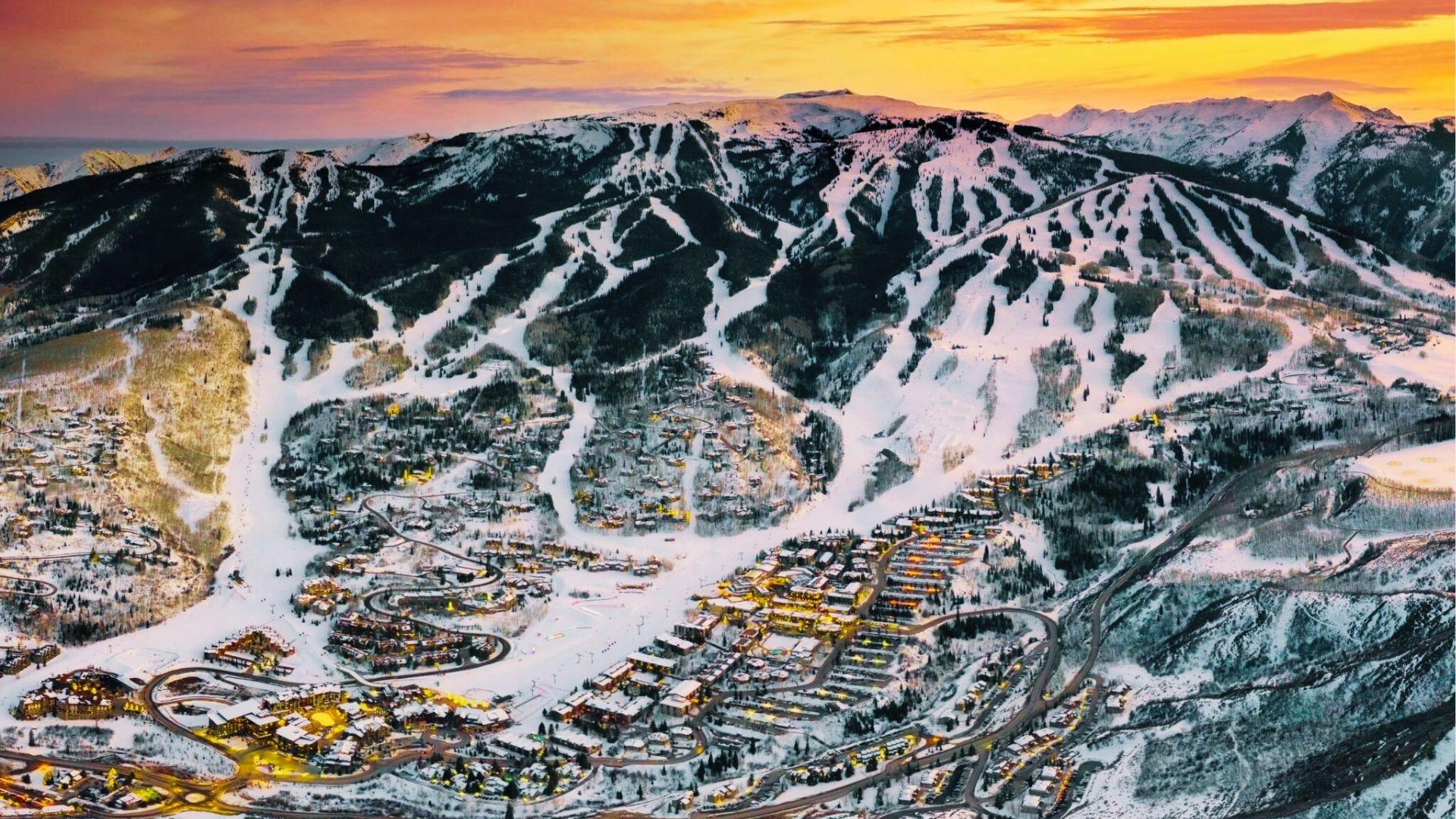 Aerial view of snow covered ski resort village at sunset with lit lodges and ski runs