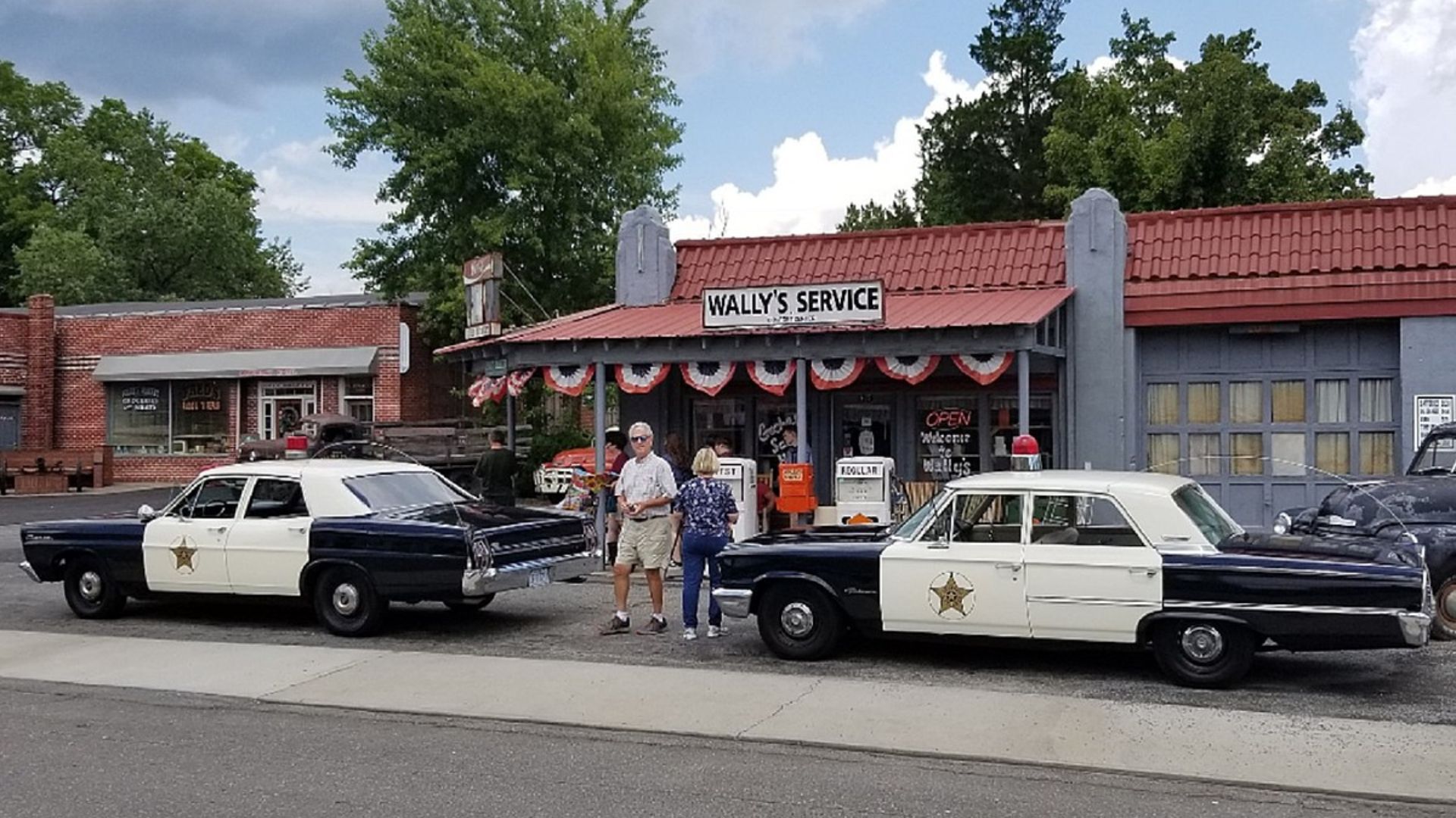 Two vintage Mayberry sheriff patrol cars parked outside Wally’s Service gas station replica with visitors nearby.