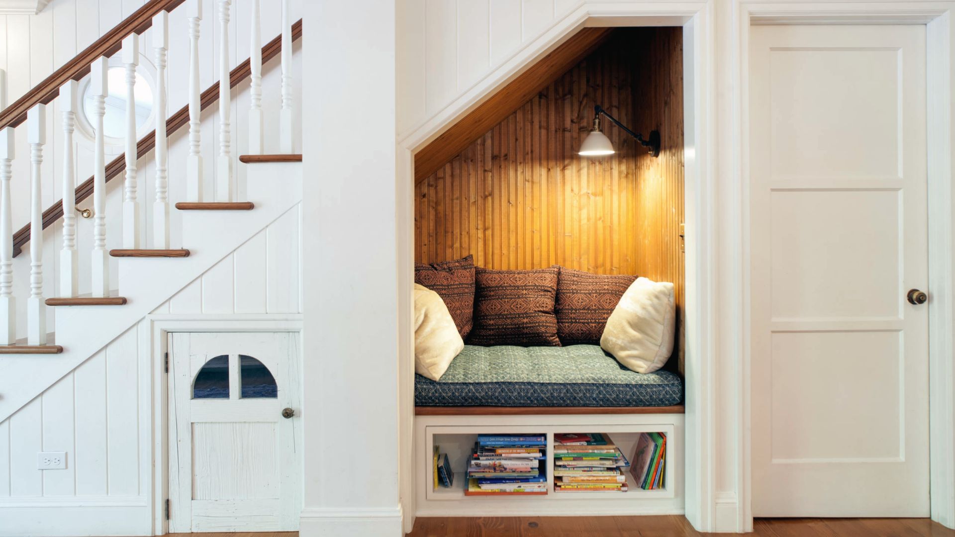 Compact reading and movie nook built under a white staircase, featuring wood paneling and a cushioned bench with pillows.