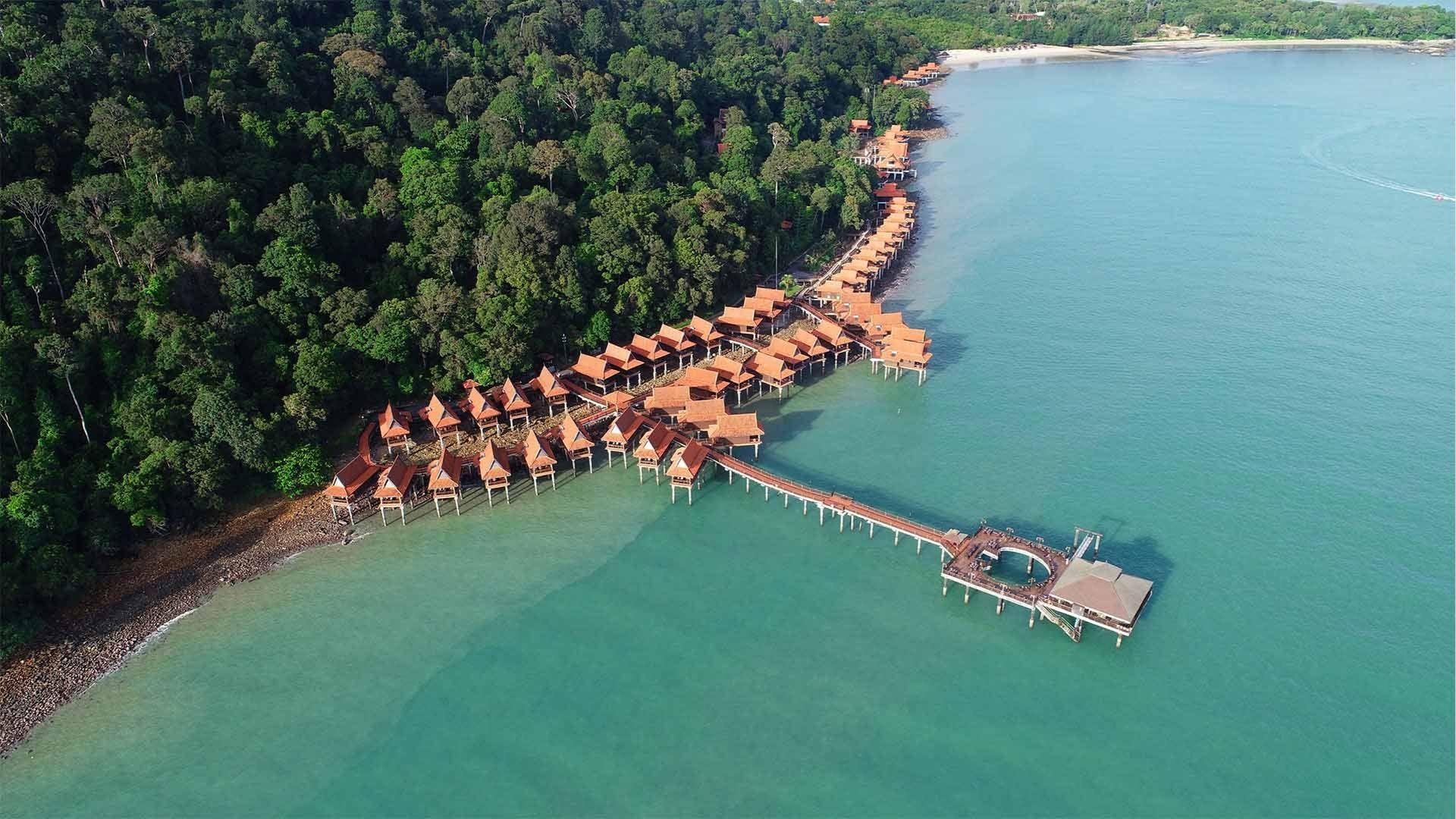 Aerial of red roofed overwater villas connected by pier extending into turquoise sea by forest