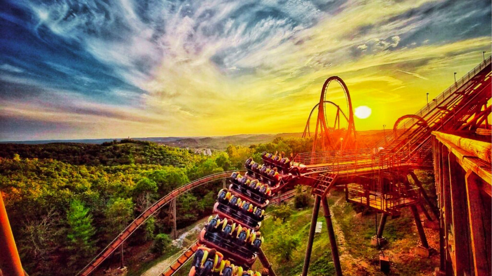 Roller coaster full of riders plunges down track at sunset with loops and forest below