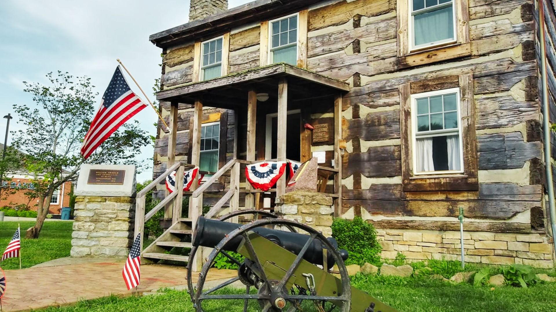 Historic William Arnold Log House decorated with American flags and a cannon out front in Williamstown, Kentucky.