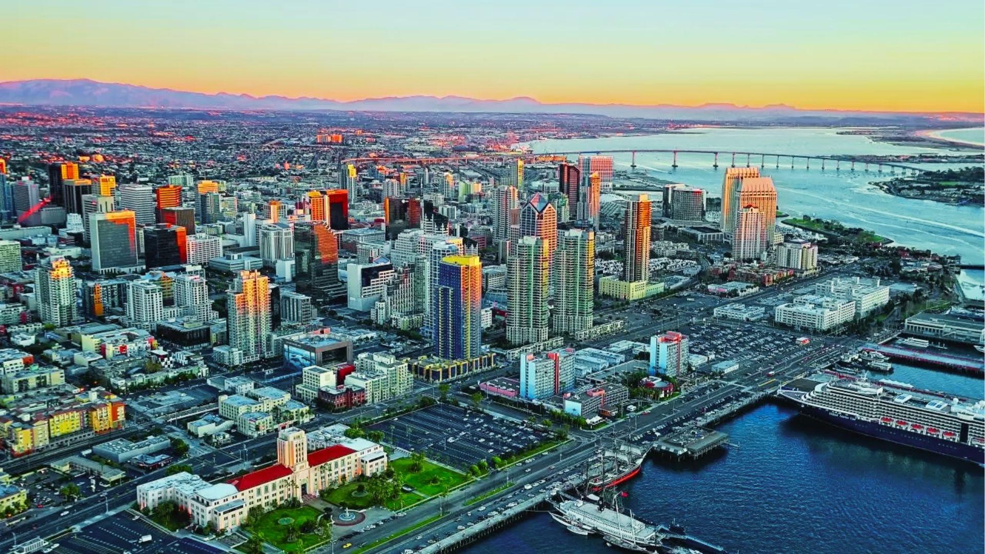 Aerial view of downtown city skyline at sunset with bridge, harbor, cruise ship, and distant mountains