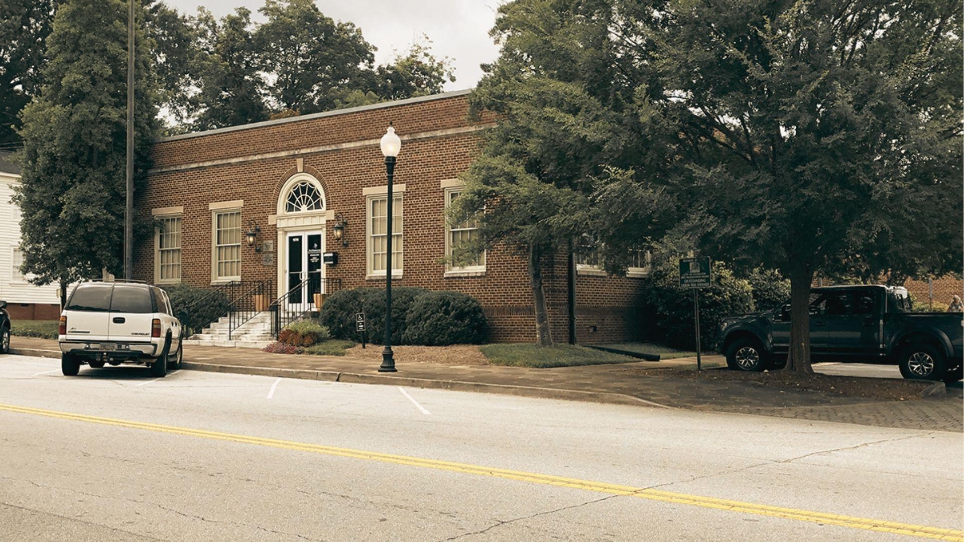 A red brick municipal building with arched doorway and white trim, flanked by trucks on a quiet street.