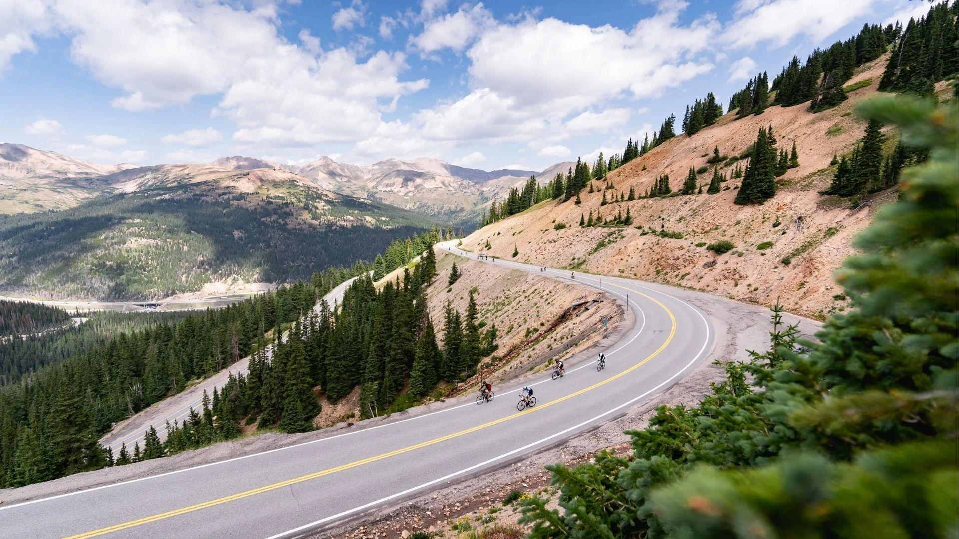 Cyclists ride up winding mountain road through pine forest with distant peaks under cloudy sky