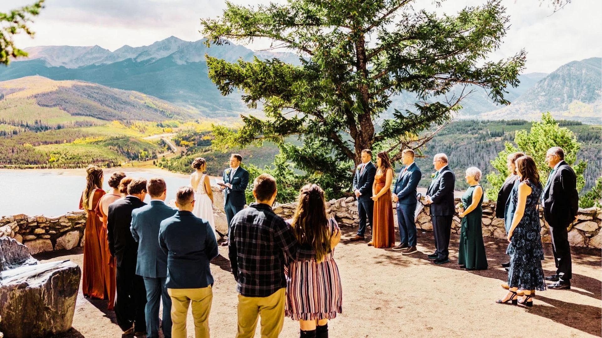 Outdoor mountain wedding ceremony with guests, bride, groom, lake, autumn trees, and peaks behind