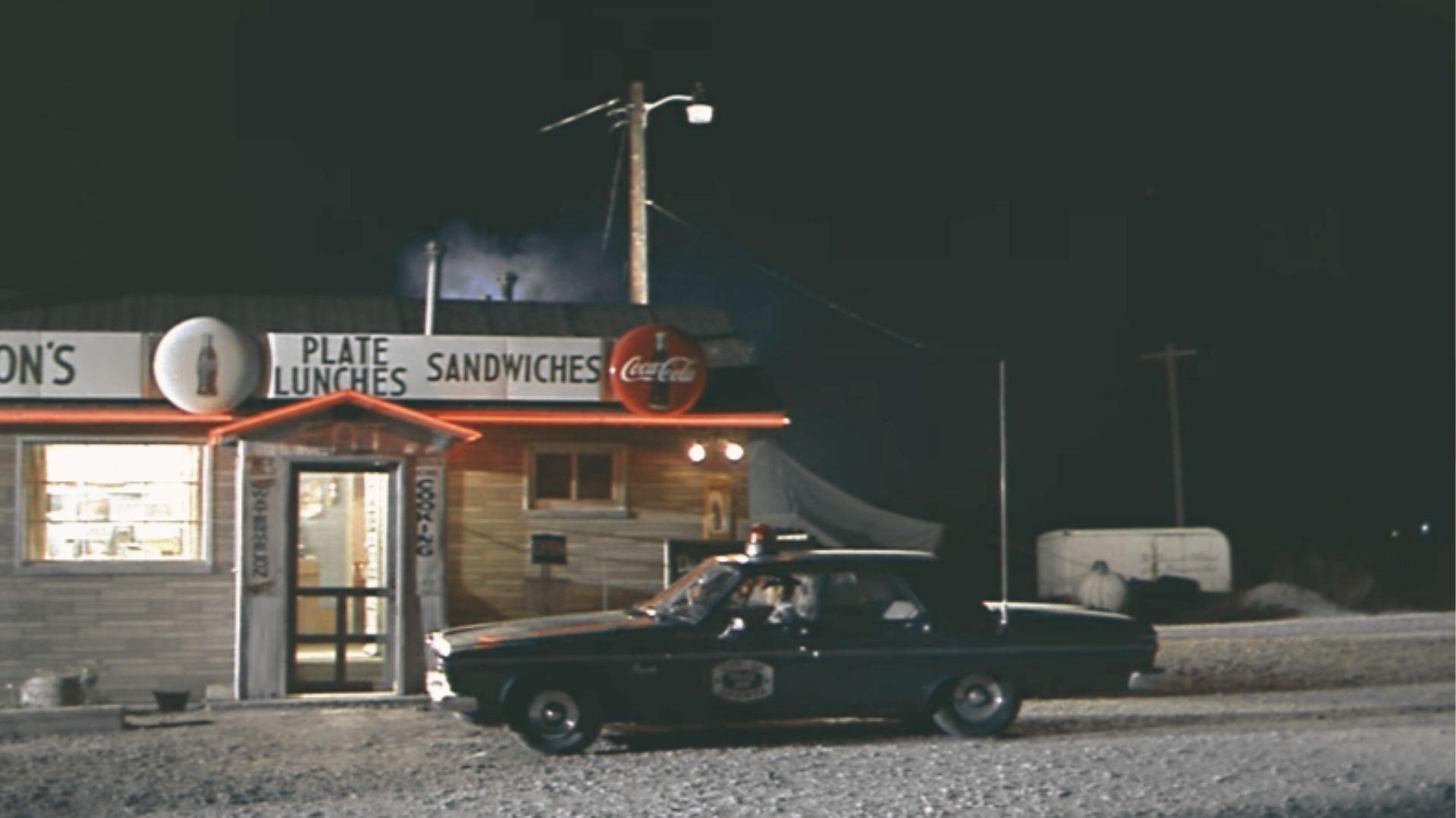 A police car idles at night outside a diner with neon Coca-Cola signs and “Plate Lunches Sandwiches” lettering.