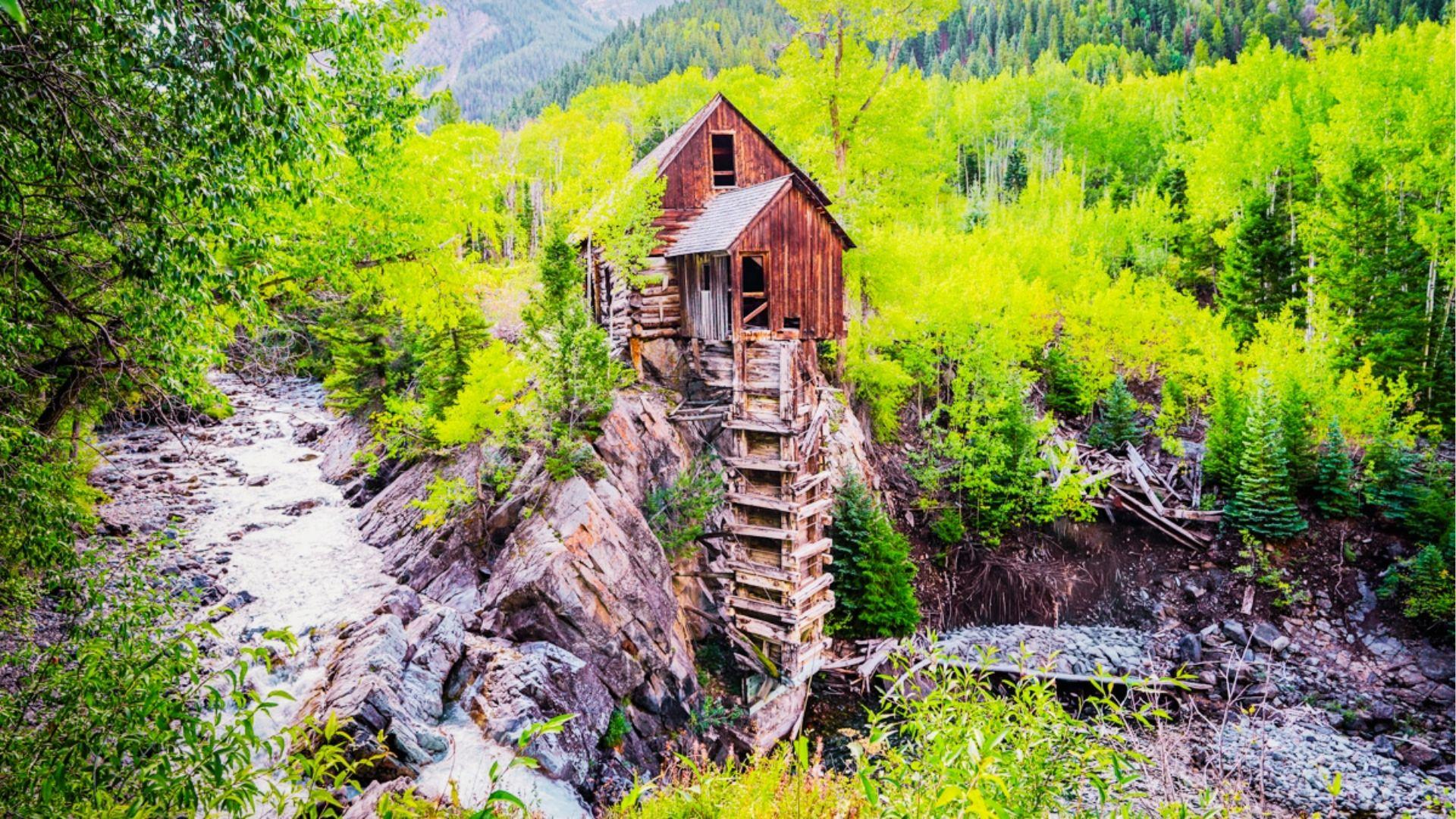 Historic wooden Crystal Mill perches on rocky cliff beside rushing stream in aspen forest