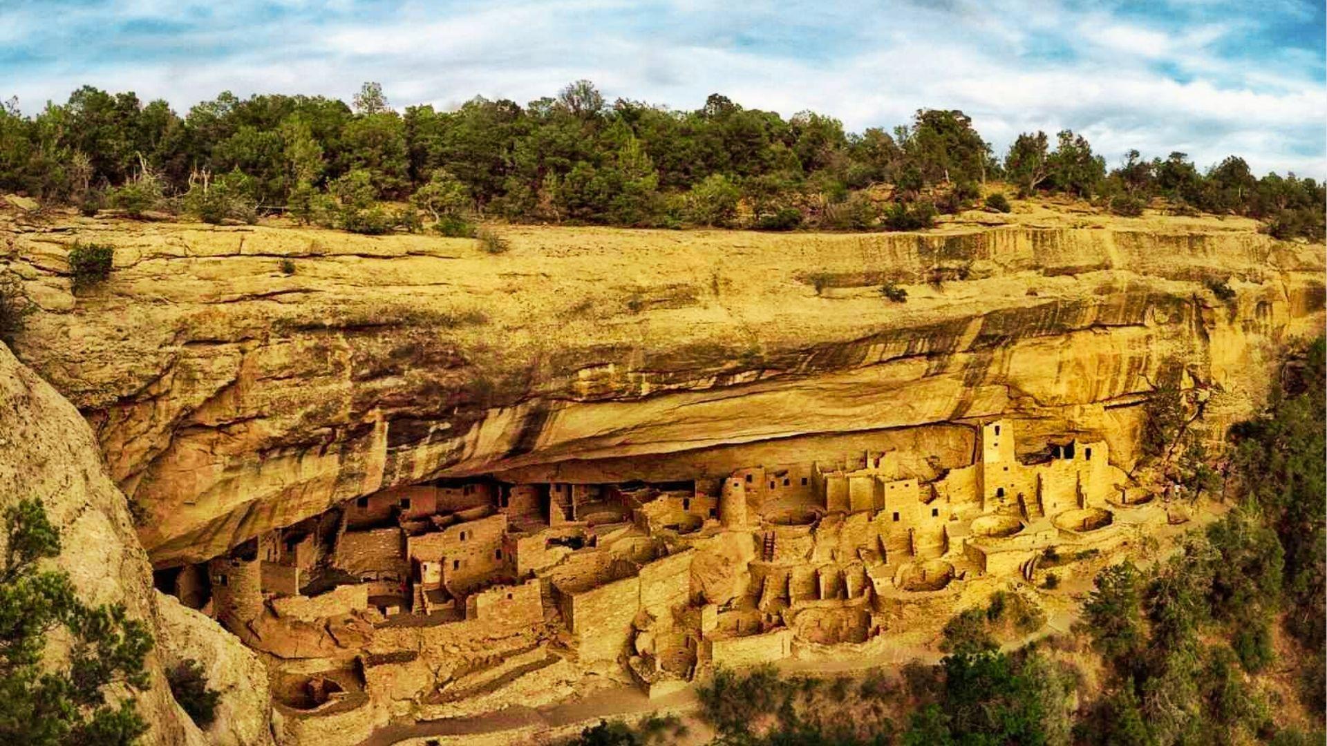 Ancient cliff dwellings built into sandstone alcove surrounded by green trees under blue sky