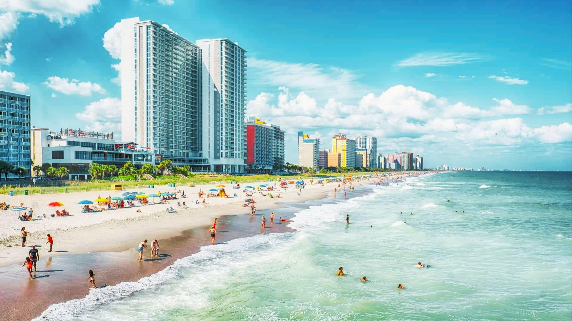 Crowded beach with colorful umbrellas, high-rise hotels, white sand, and turquoise ocean under blue sky