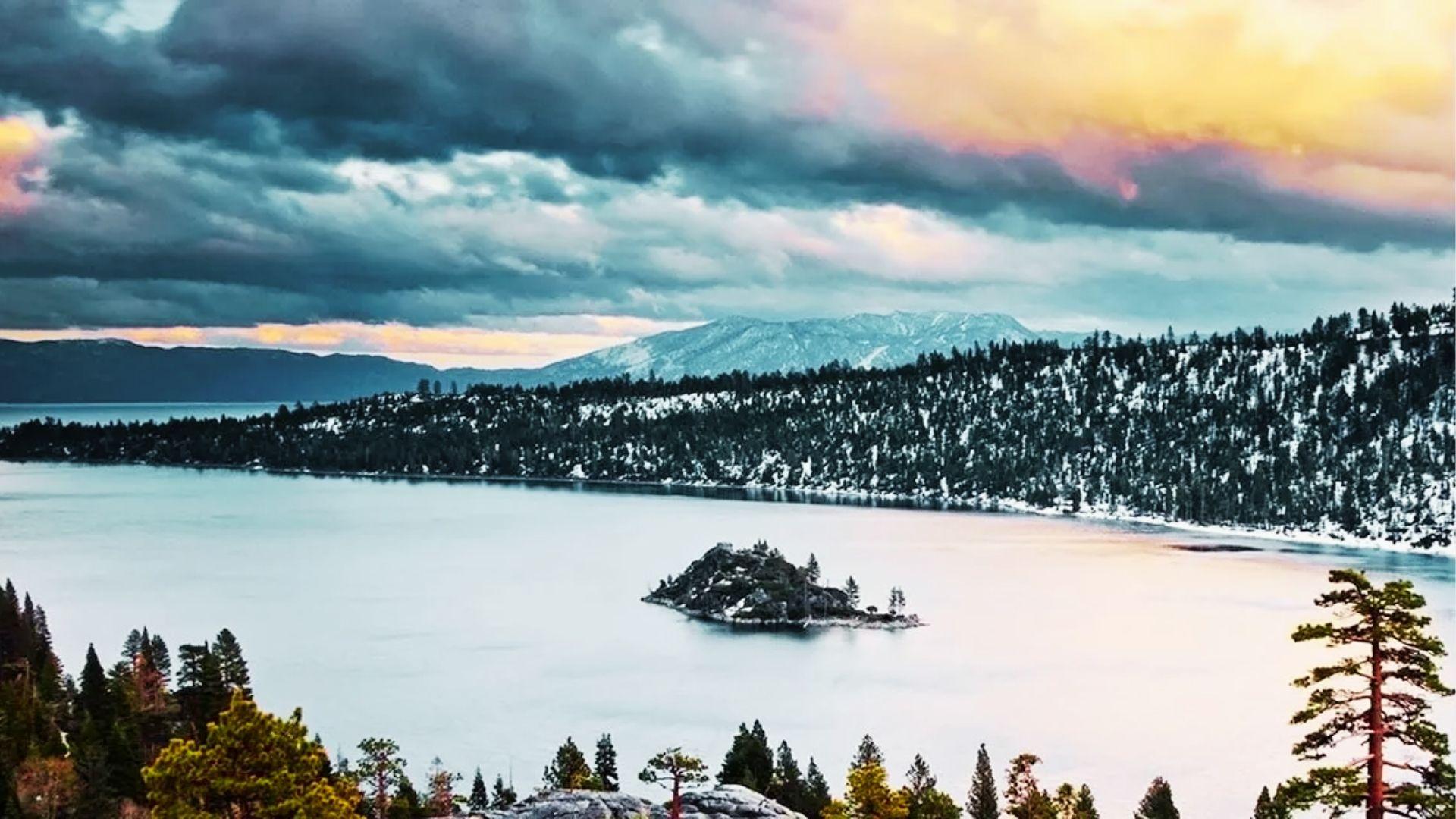 Snowy alpine lake with small rocky island, pine forests, mountains, and dramatic sunset clouds