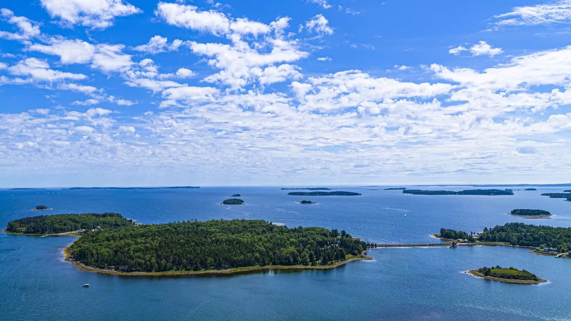 Wide aerial shot of Oak Island under blue sky, connected by causeway, surrounded by islets.