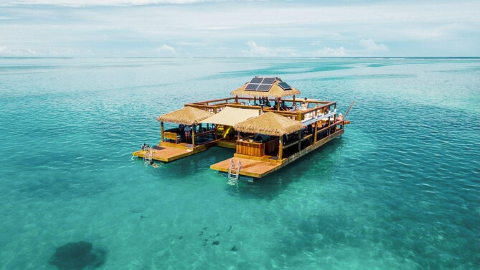 Floating thatched roof bar with solar panels sits on clear turquoise ocean under blue sky
