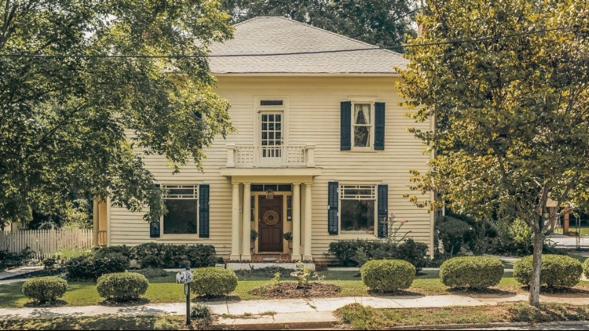 A yellow two-story house with black shutters and a columned porch sits behind manicured shrubs.