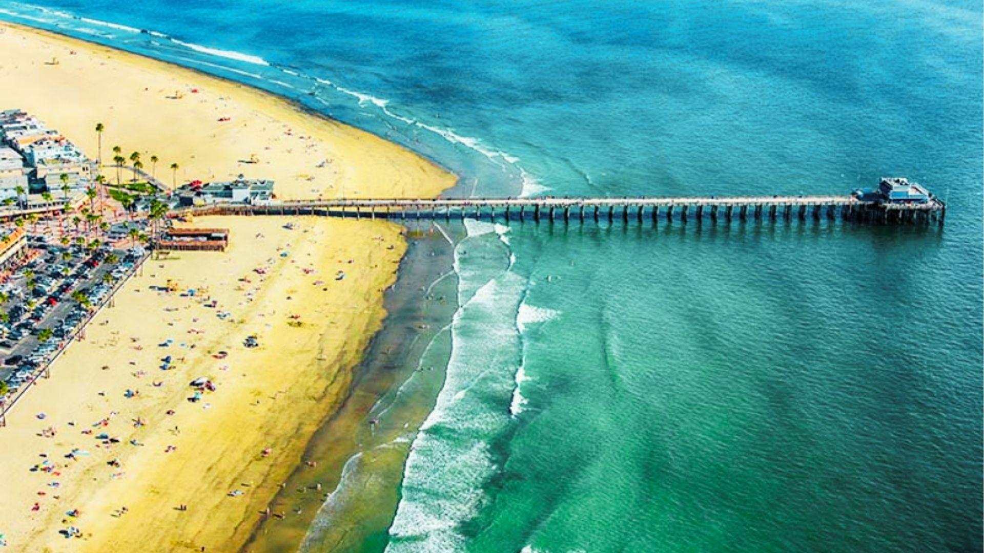 Aerial view of long beach pier extending into turquoise ocean with sand, palm trees, crowds
