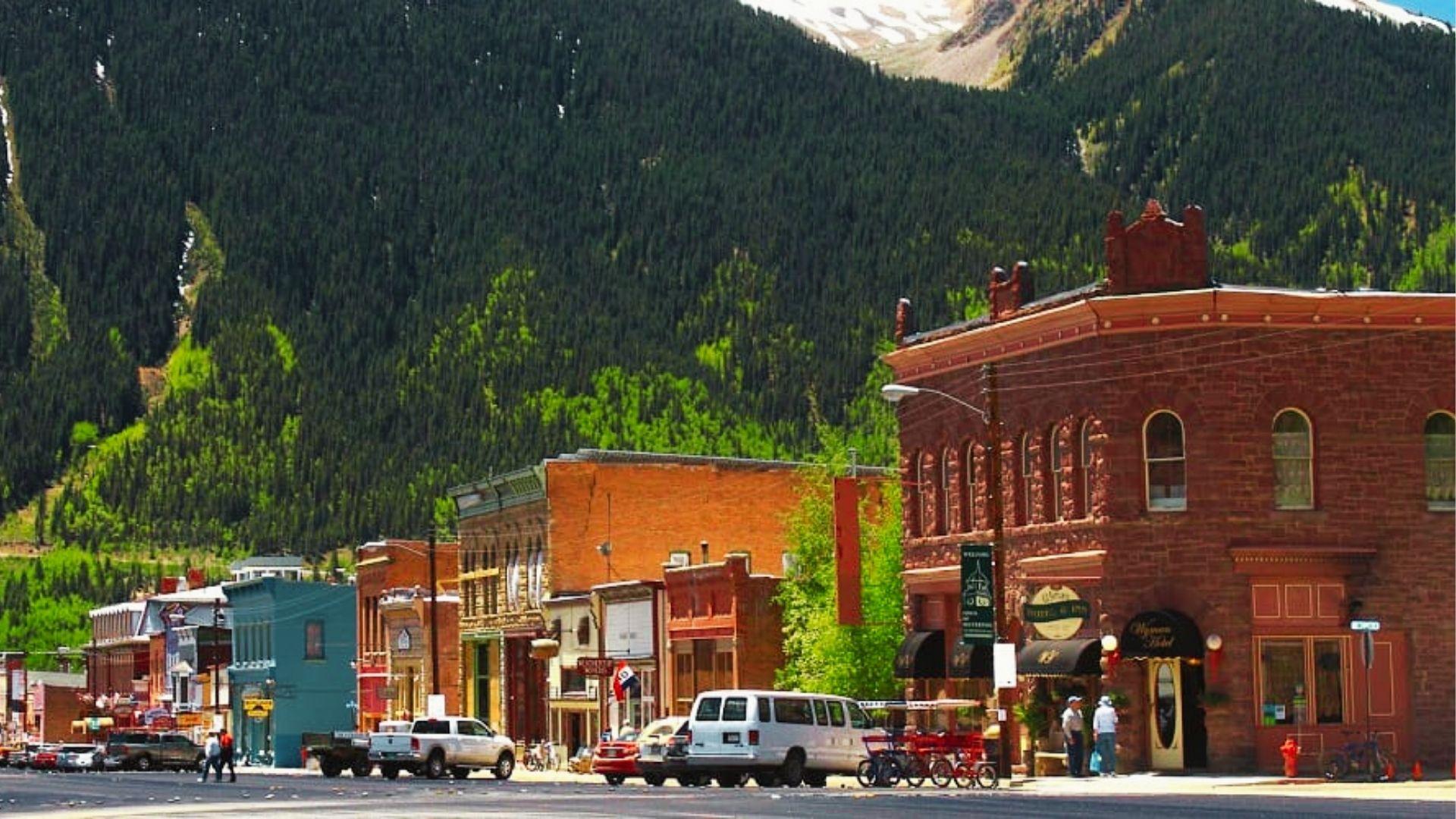 Small town street with red brick buildings, parked cars, bicycles, and green forested mountains