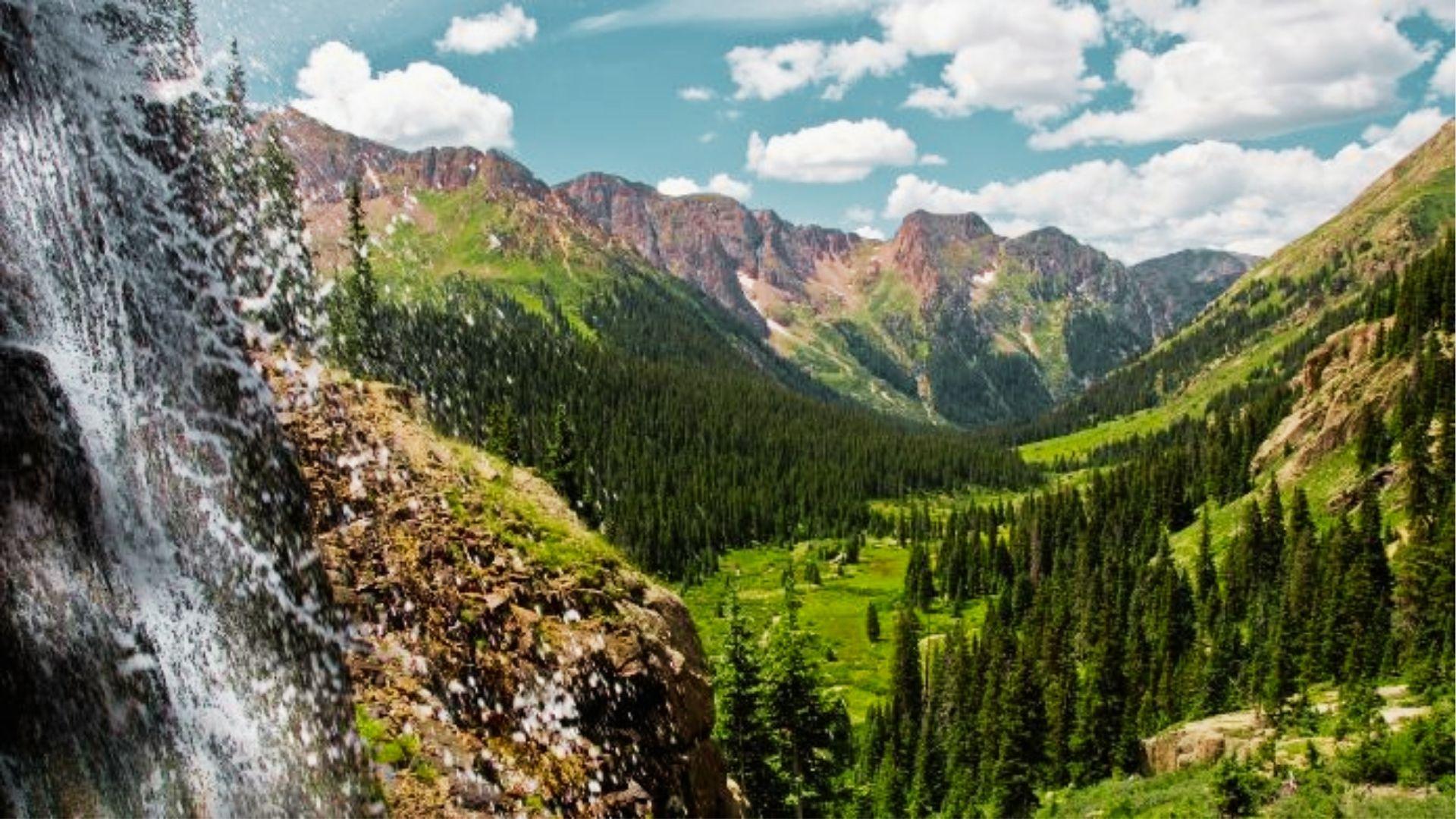 Waterfall sprays mist over green valley with pine forest and rugged mountain peaks beyond