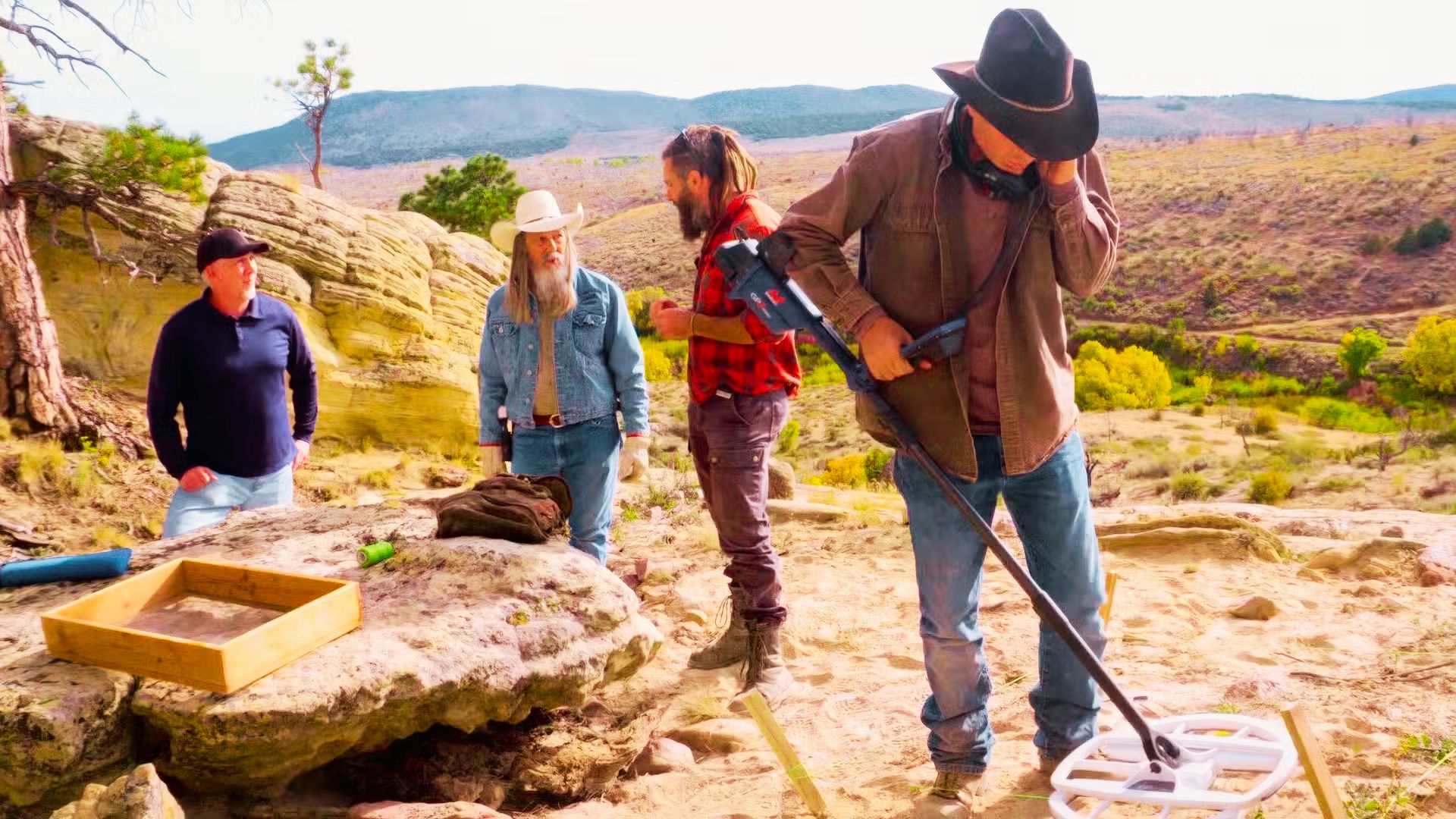 A man in a cowboy hat uses a large metal detector on rocky terrain at Blind Frog Ranch while three others watch.