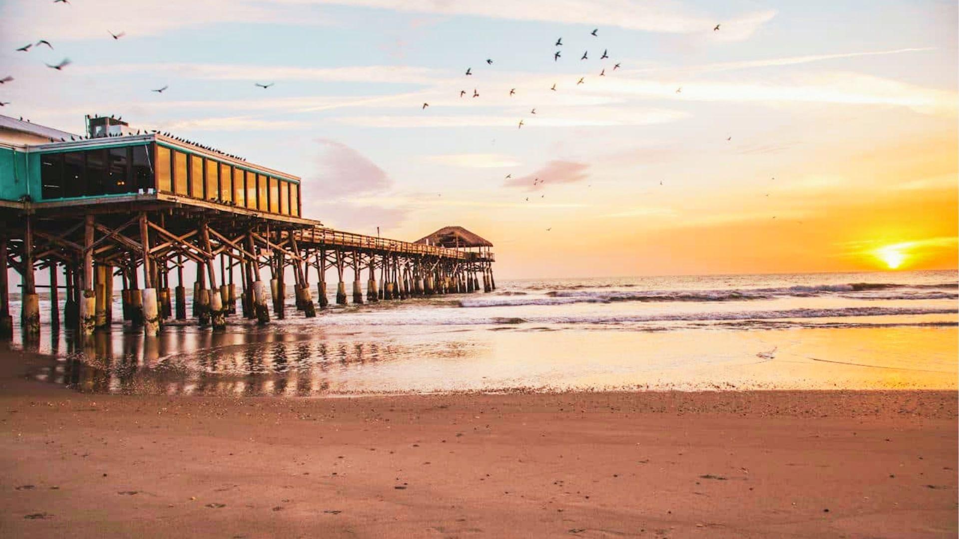 Wooden pier with restaurant extends over ocean waves at golden sunrise with birds flying overhead
