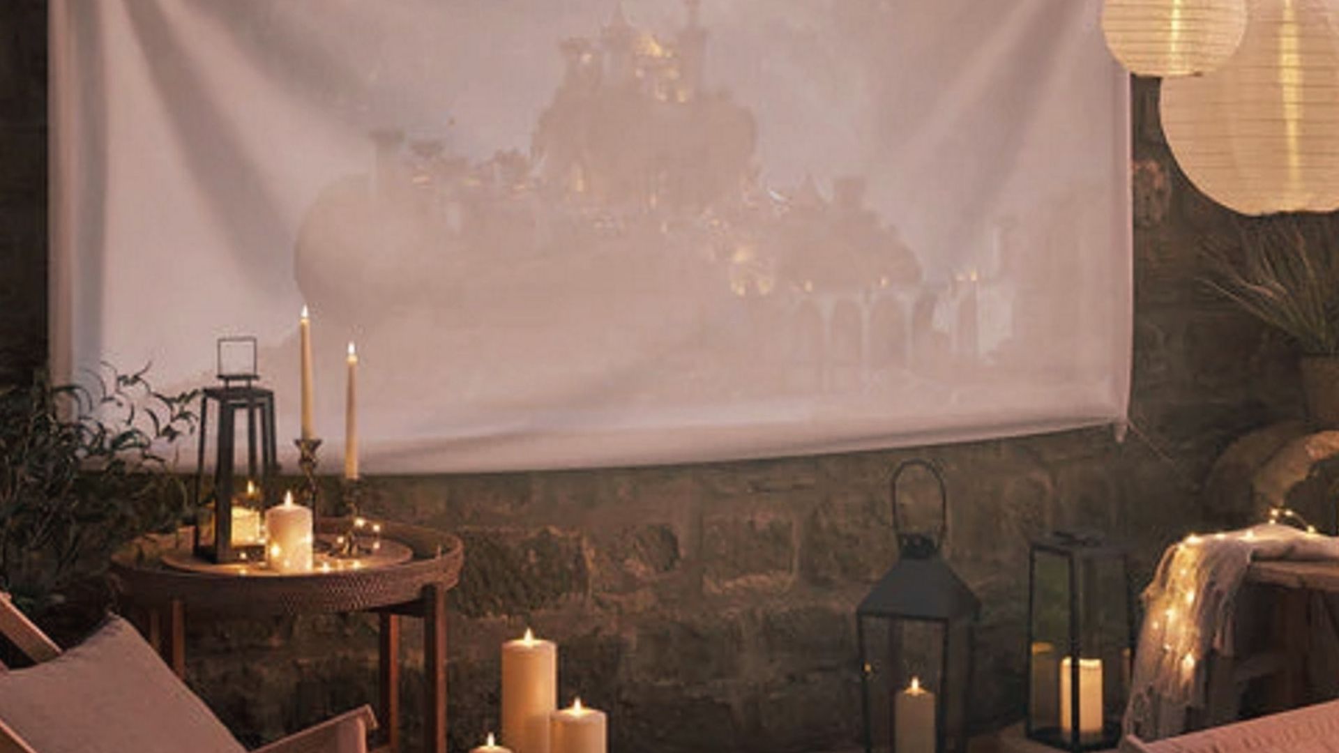 The outdoor projector screen hangs on a stone wall, glowing with a castle scene, surrounded by lanterns and candles.