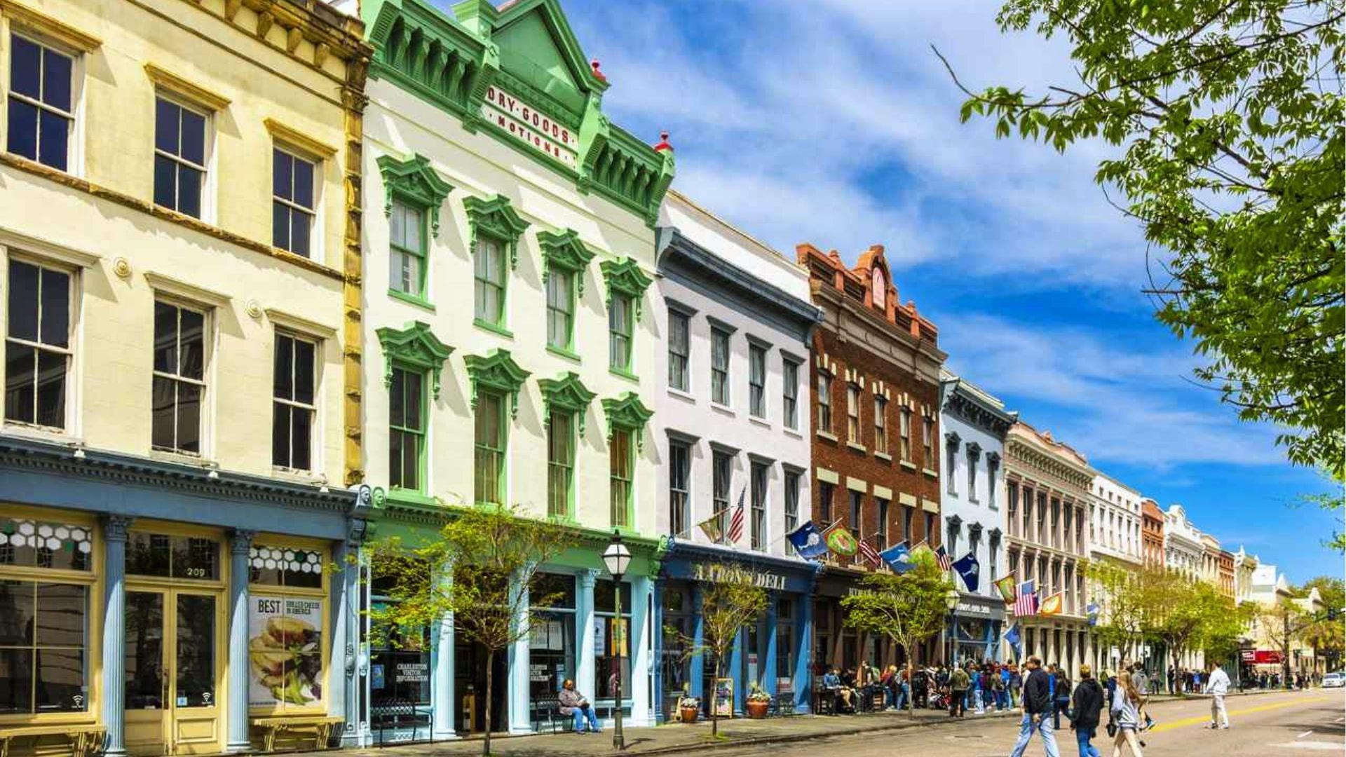 Colorful historic buildings with wrought-iron details and flags line a sunny street with crowds strolling.