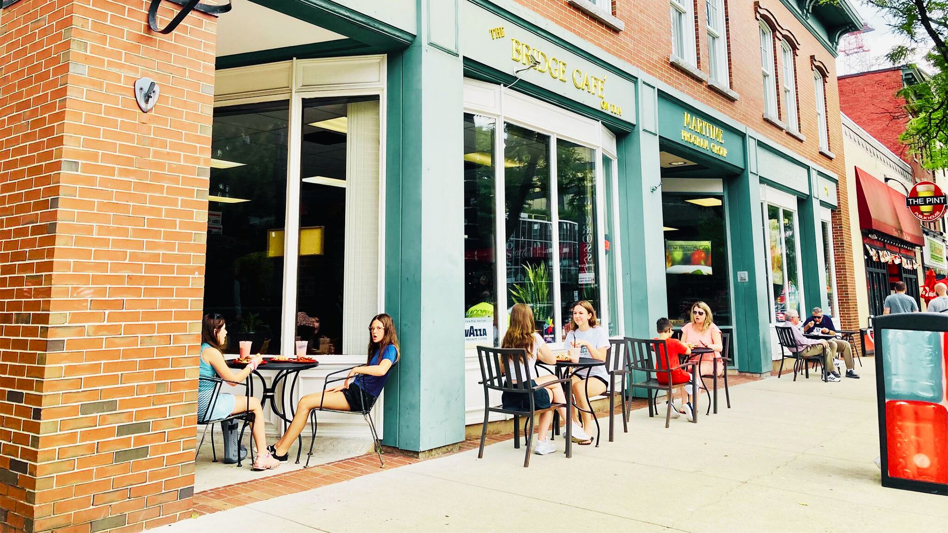 People sit at outdoor café tables along a brick storefront on a sunny downtown street in Manchester-by-the-Sea.