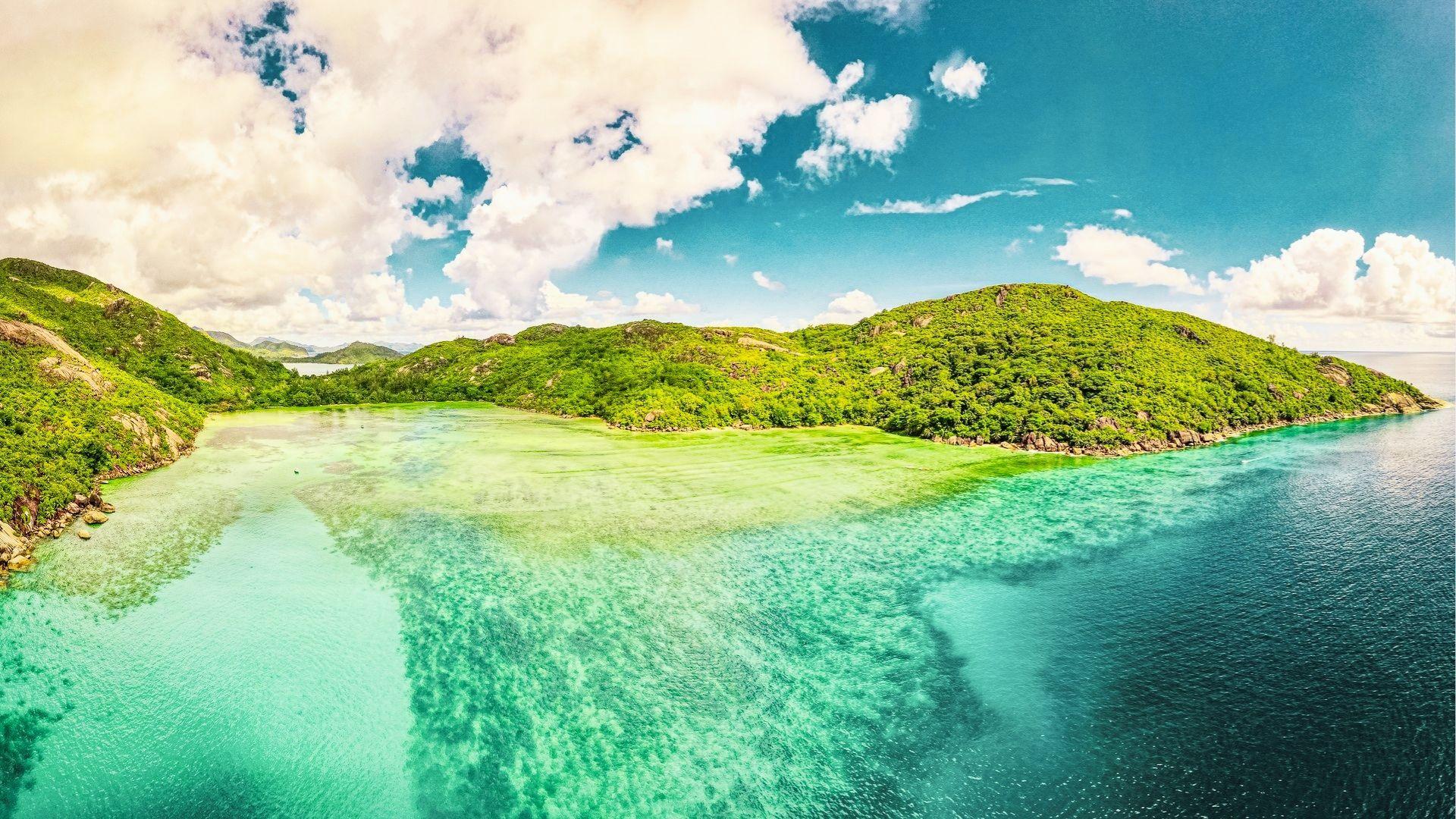 Aerial of lush green hills meeting clear turquoise bay with shallow reef under blue clouds