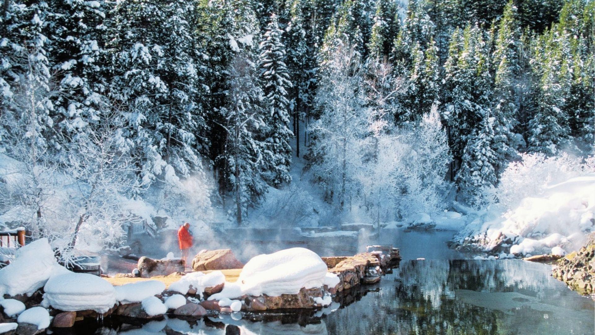 Person in red robe stands by steaming hot springs surrounded by snow-covered pine forest