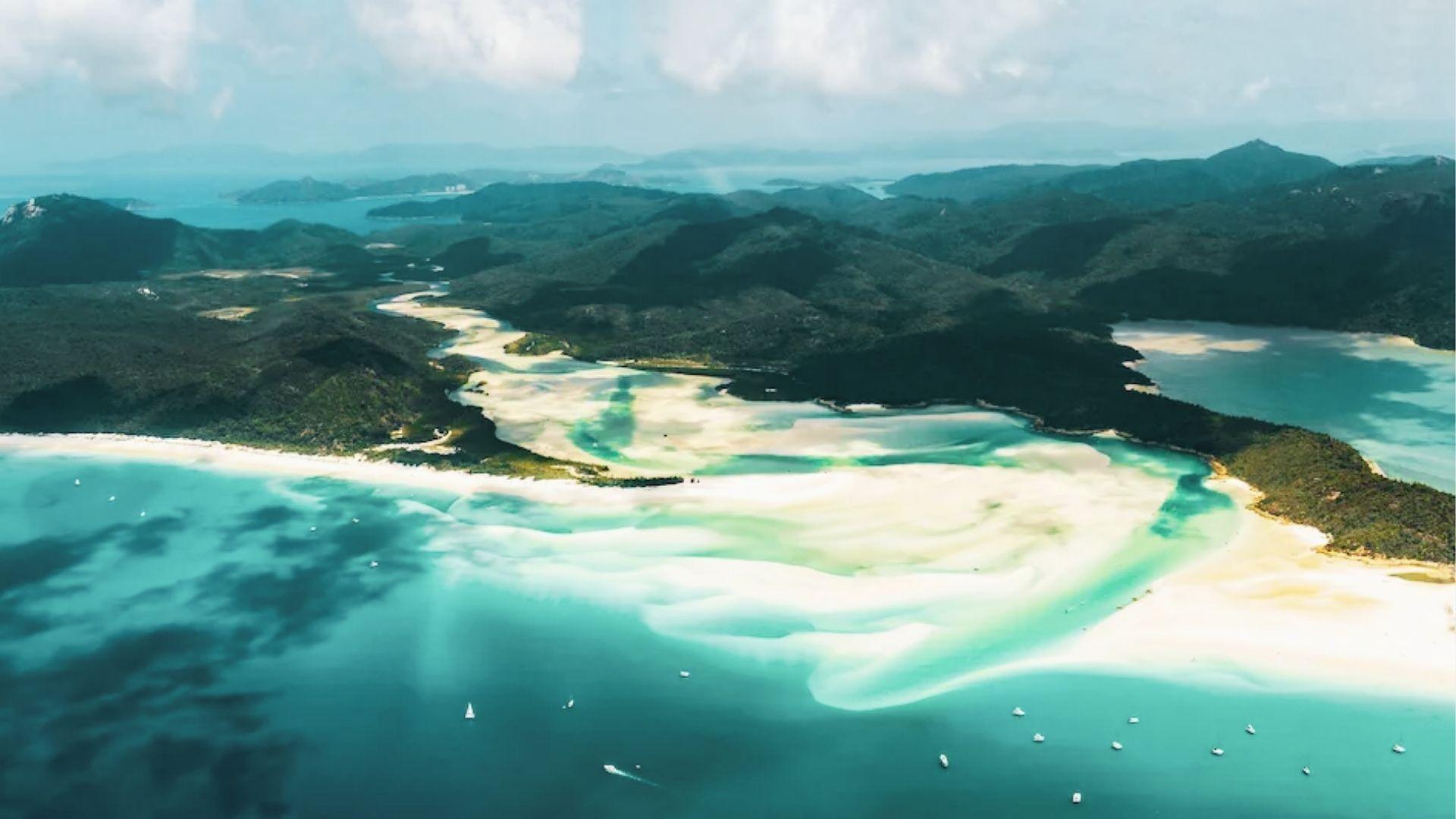 Aerial of winding white sand beach with turquoise water, boats, and forested hills behind
