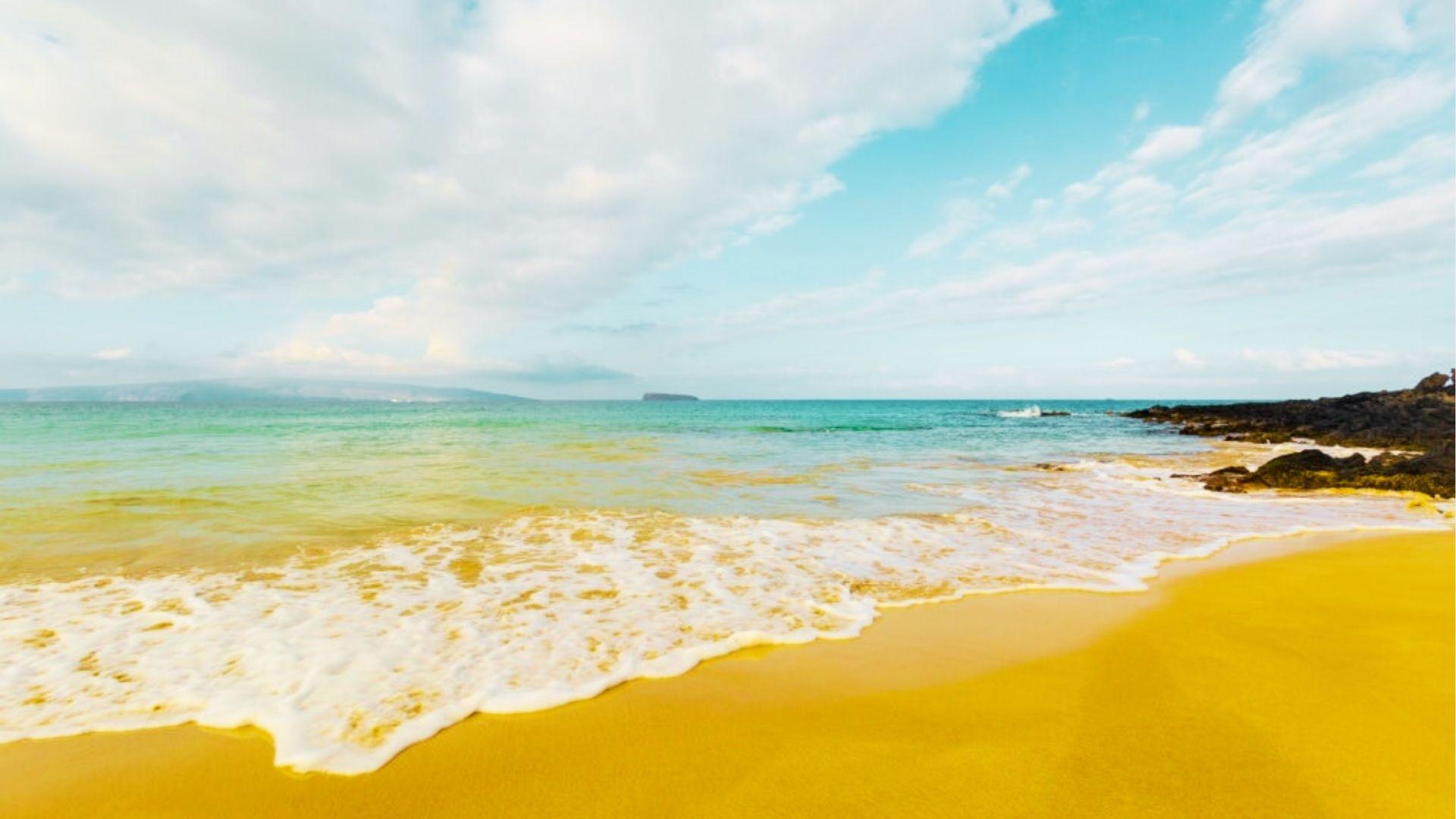 Golden sand beach with gentle waves, black rocks, turquoise water, and distant island horizon sky