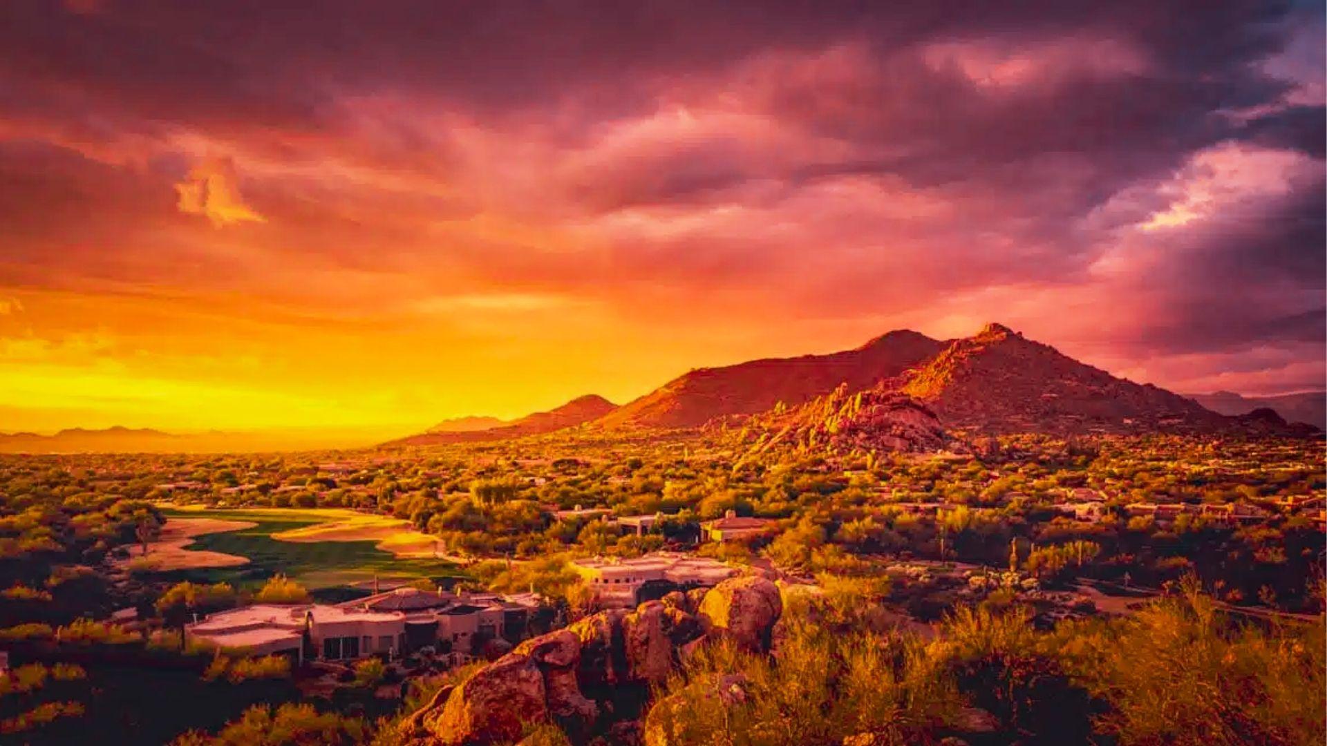 Desert golf course and homes glow under dramatic orange sunset with rugged mountains and stormy sky