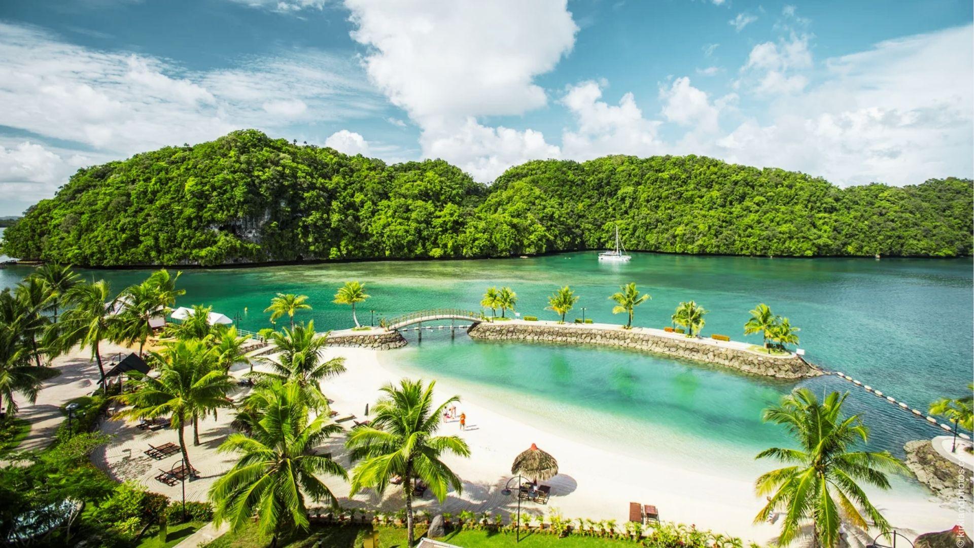 Tropical beach resort with palm trees, white sand, bridge to island, sailboat, and hills