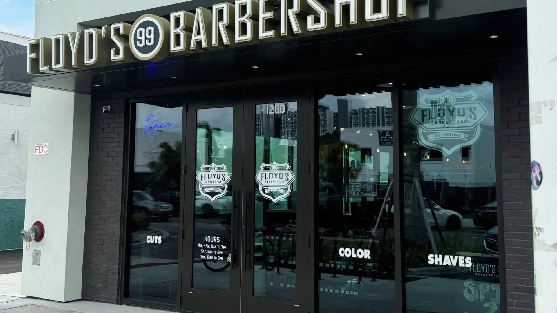 Modern black brick storefront with neon “Open” sign and glass doors advertising cuts, color, and shaves.