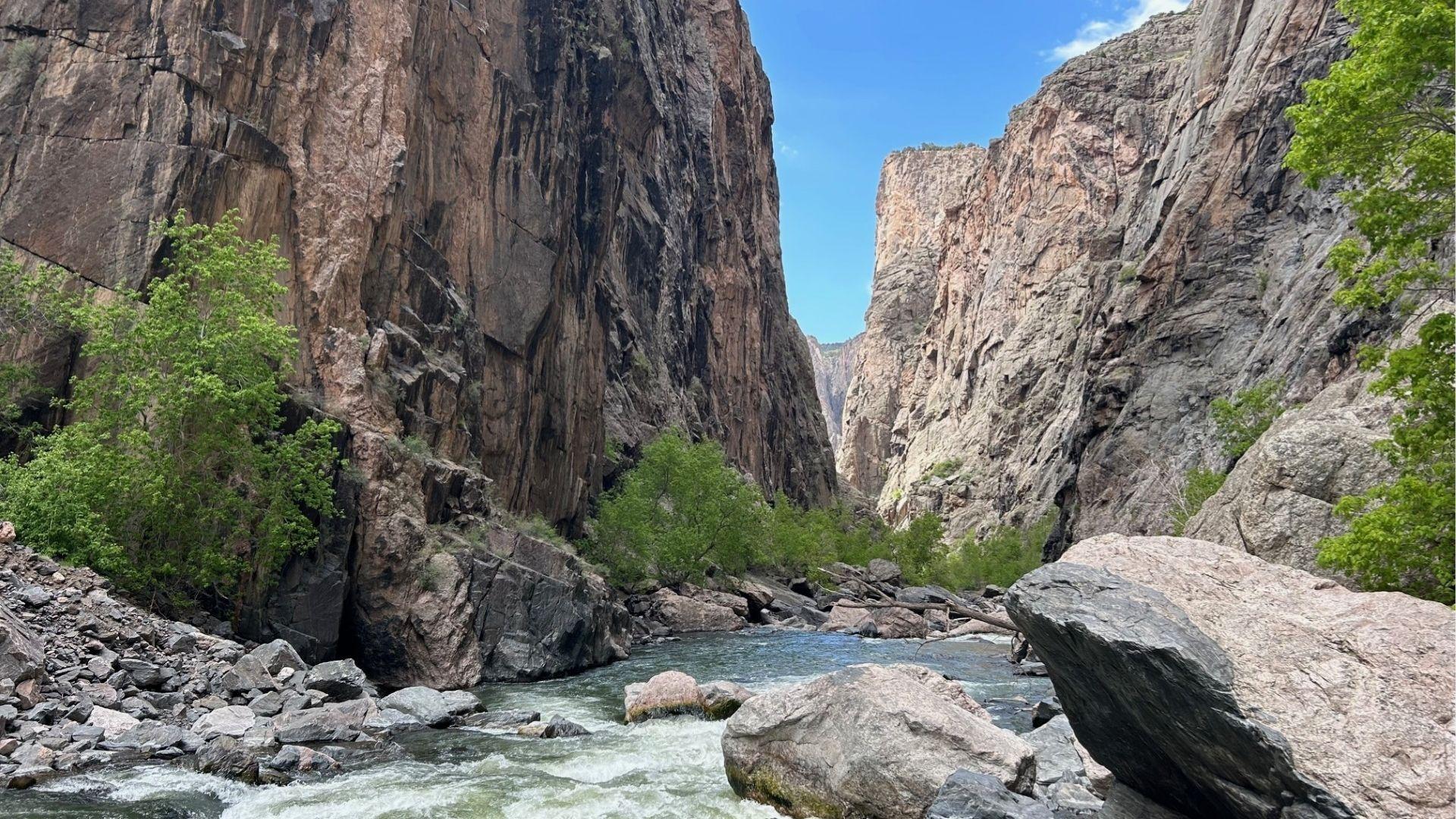 Deep rocky canyon with steep cliffs, flowing river, boulders, and green trees under clear sky