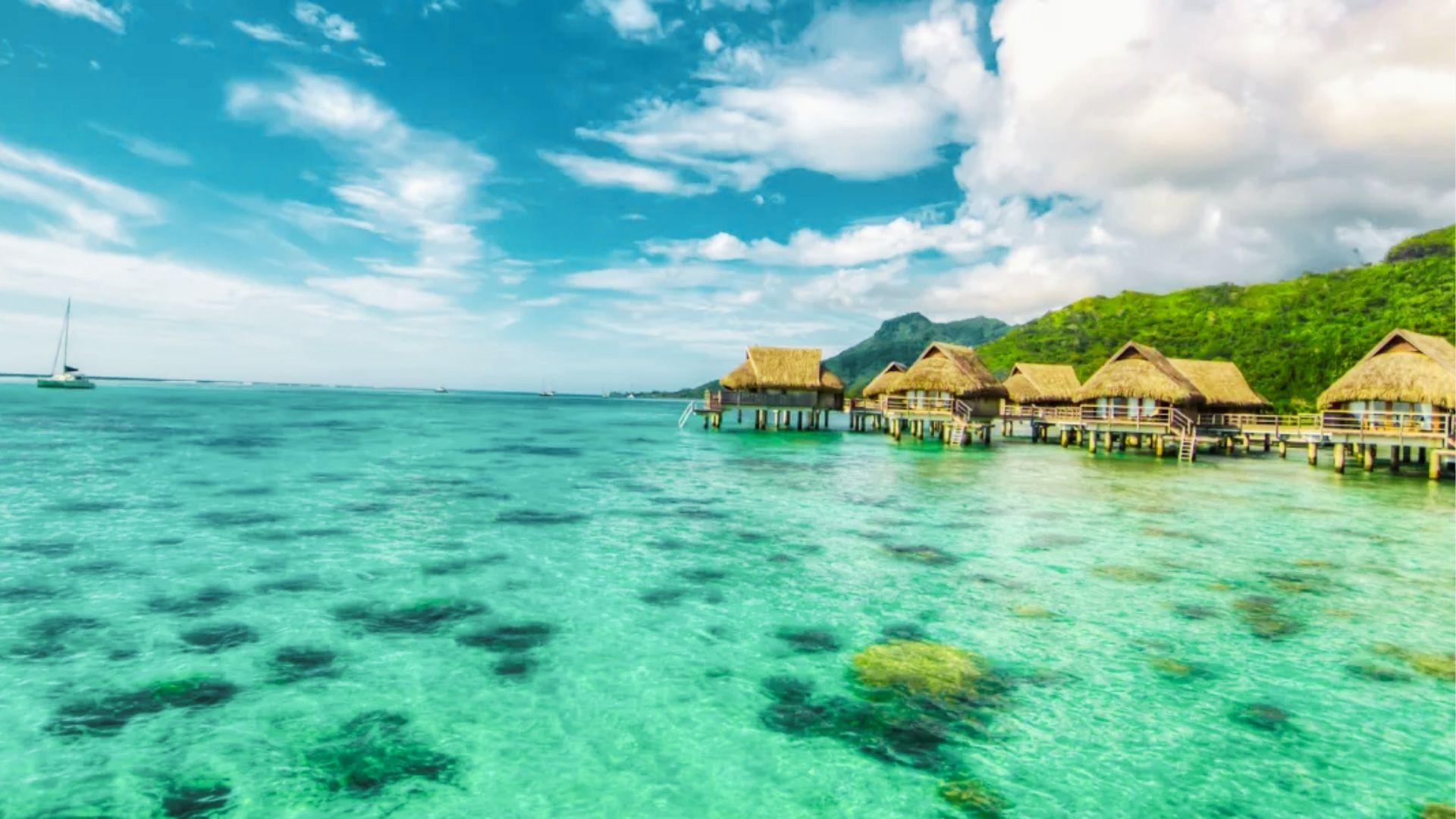 Thatched overwater bungalows on stilts extend into clear turquoise lagoon with green mountains beyond