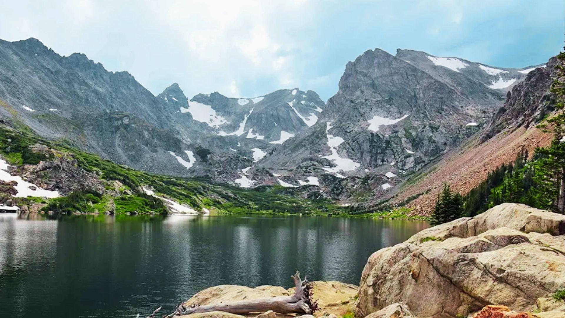 Clear alpine lake surrounded by snow patched peaks, pine forest, boulders, and fallen log foreground