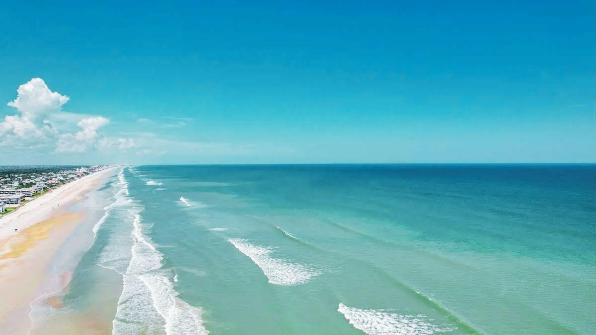 Turquoise ocean waves roll onto long sandy beach lined with homes under bright blue sky
