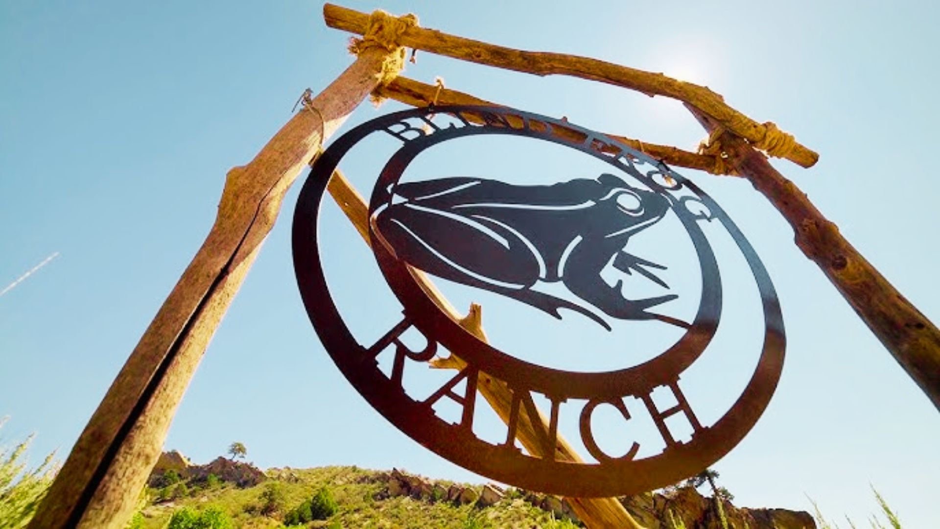 A rustic metal sign reading “Blind Frog Ranch” with a frog silhouette hangs from a wooden frame against a bright sky.