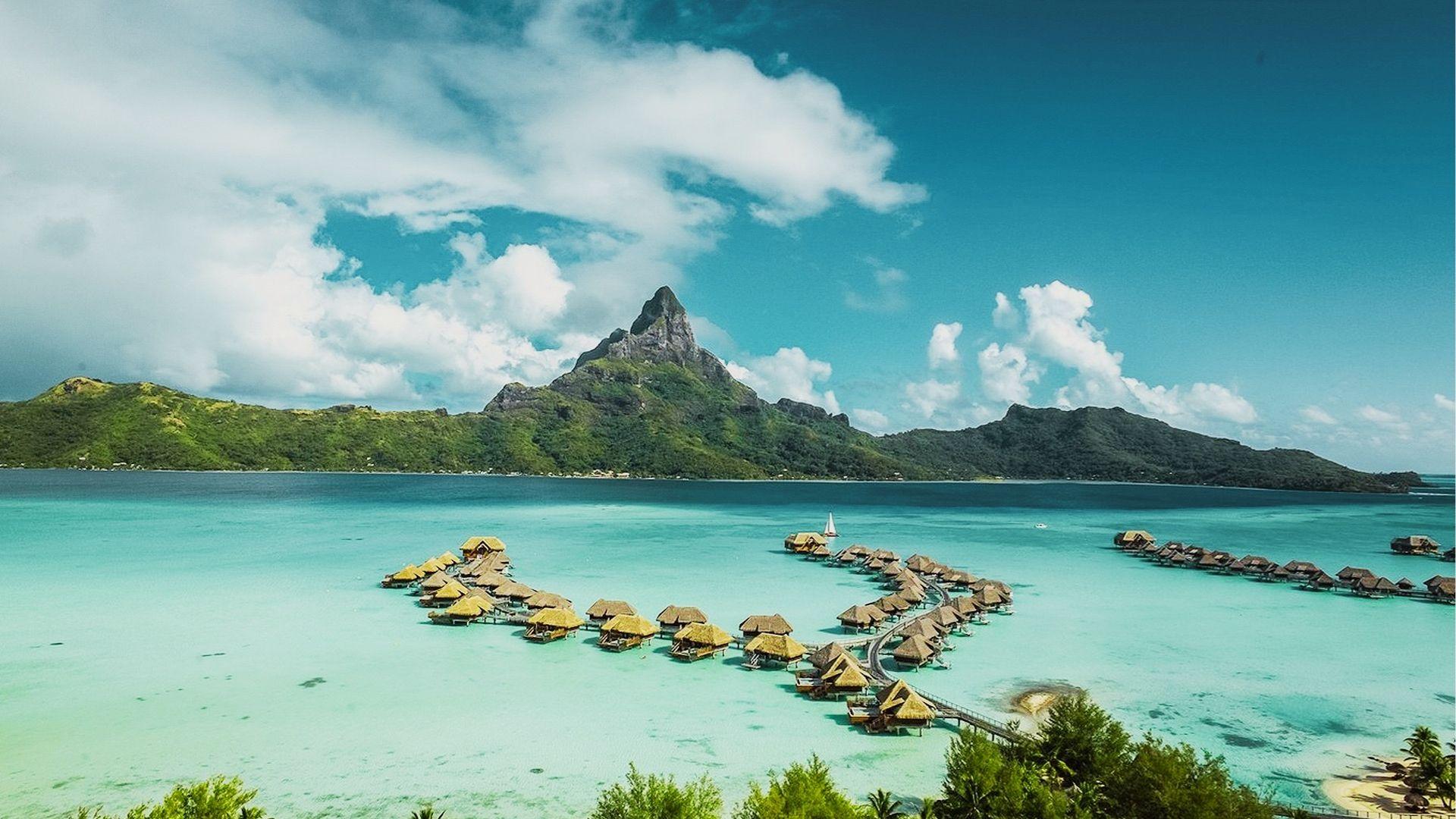 Overwater bungalows on stilts extend into turquoise lagoon with green mountain peak and distant sailboat
