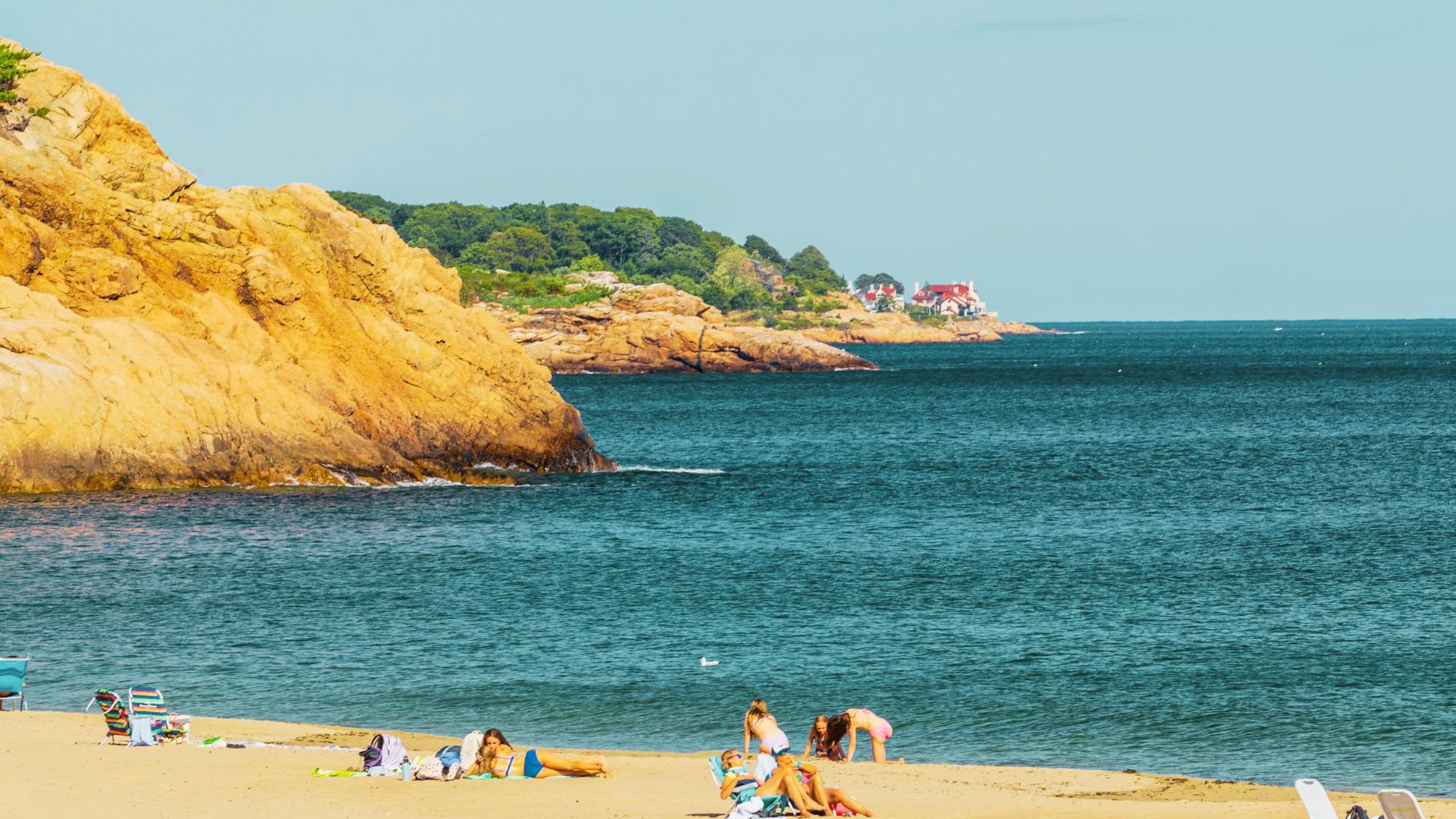 Sunbathers relax on a sandy beach in Manchester-by-the-Sea with dramatic rocky cliffs and deep blue water beyond.