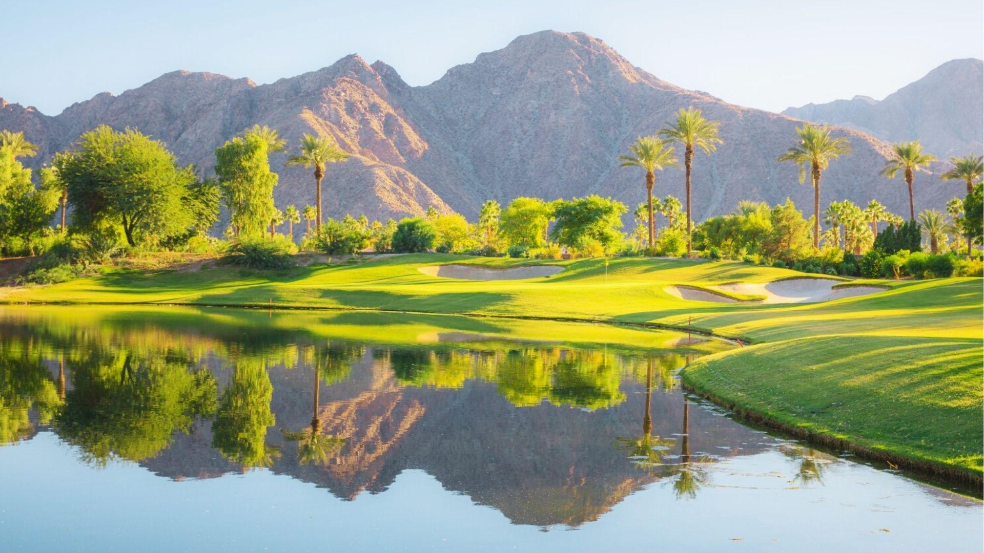 Lush green golf course with palm trees reflects in a calm lake, framed by sunlit desert mountains.