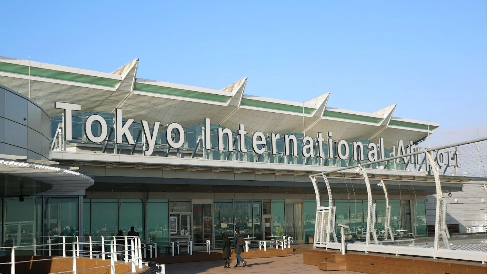 Modern exterior of Tokyo International Airport Haneda under clear blue skies with travelers walking outside.