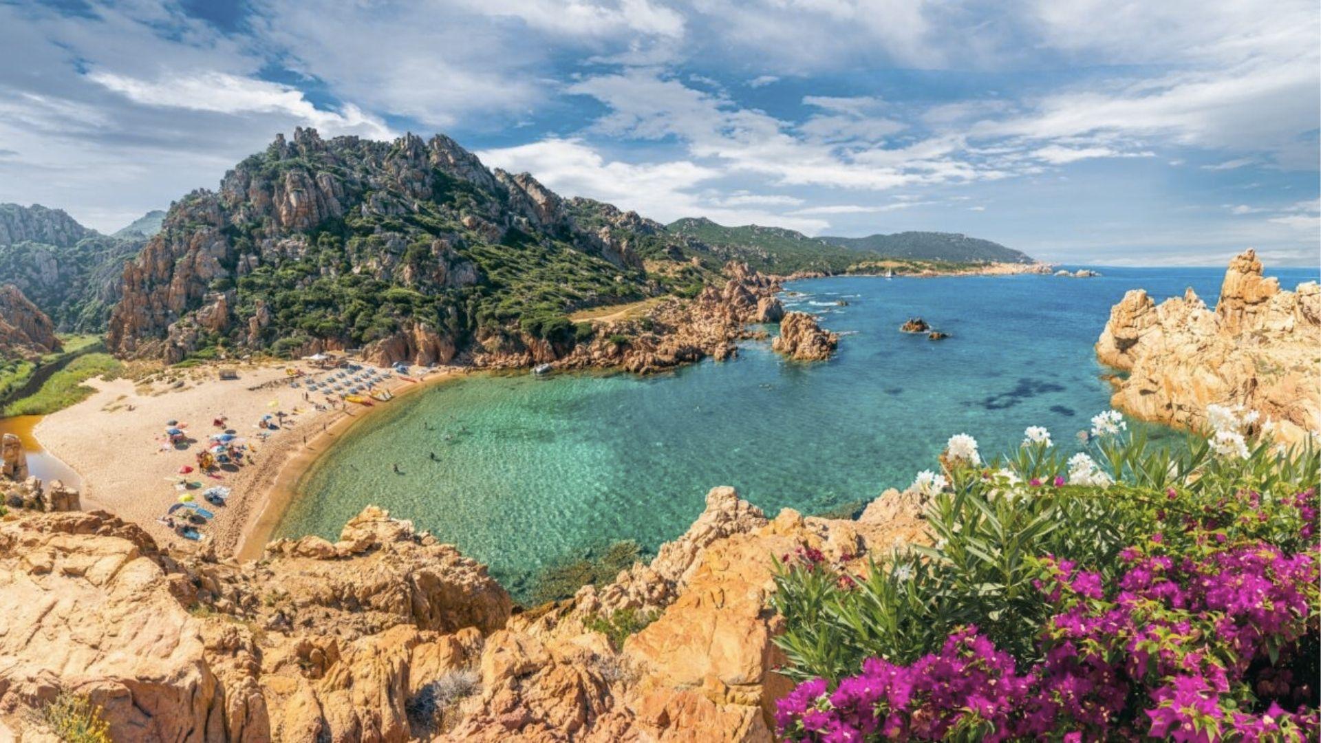 Turquoise waters lap a crescent beach framed by jagged cliffs in Sardinia, Italy.
